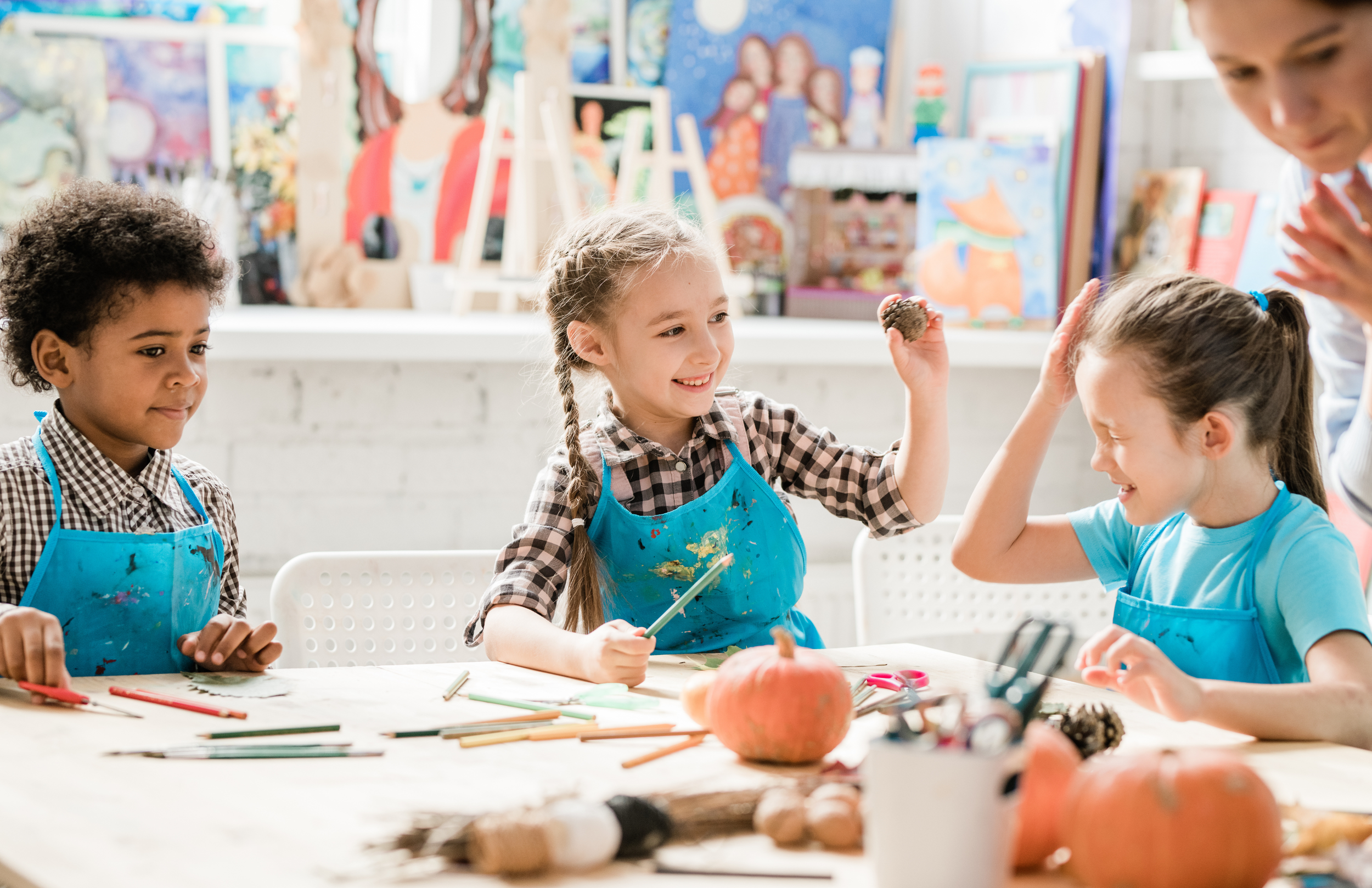 Children in a colorful art classroom engaging in a painting activity, wearing aprons, with pumpkins and art supplies on the table. Indoor playground in Howell Michigan.
