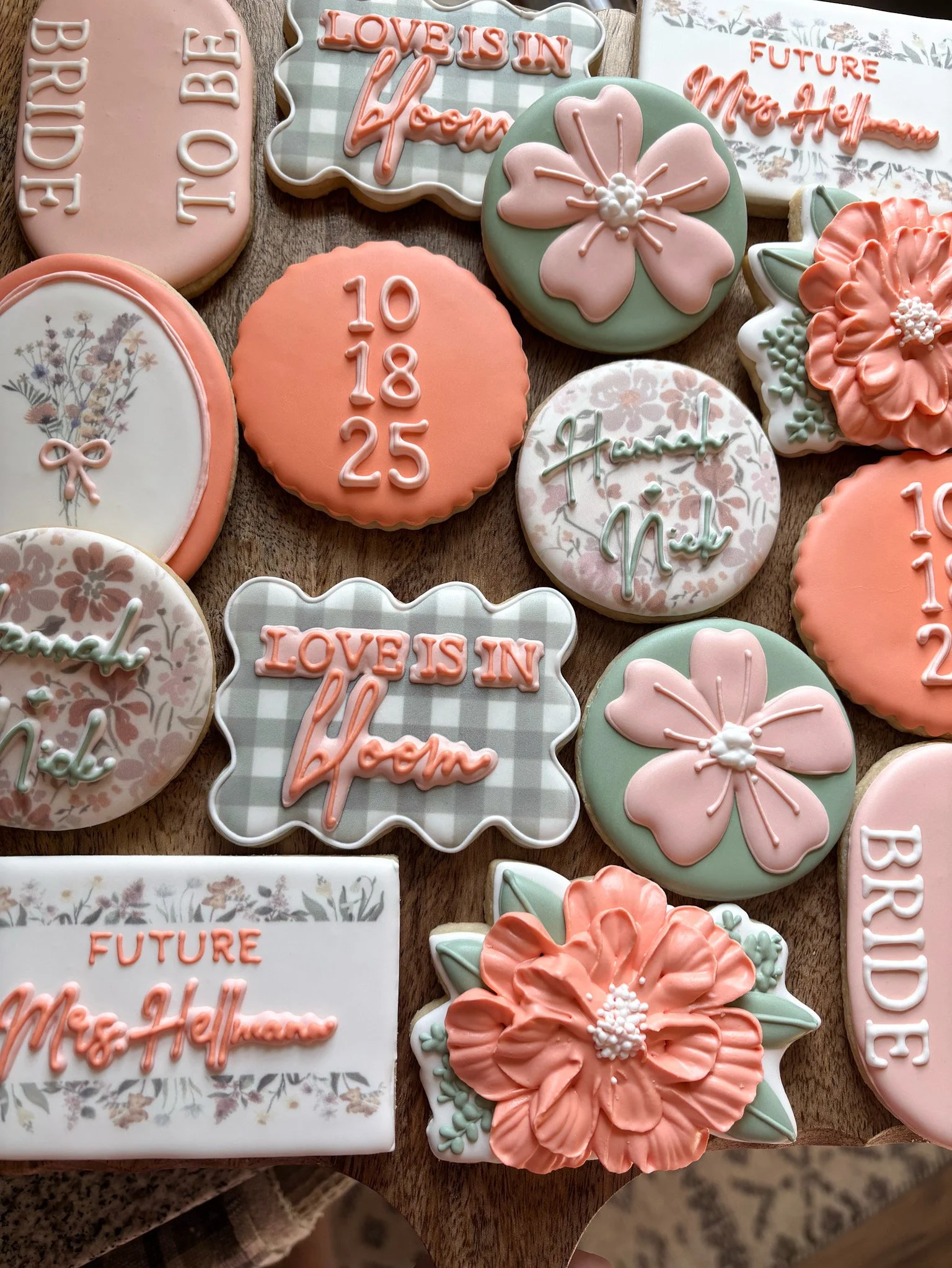 Decorative cookies with floral and text designs on a wooden surface. Some cookies have messages like 'LOVE IS IN the home,' 'FUTURE Mrs. Hellen,' and dates '10 18 25.' Others feature pink flowers, a bouquet, and floral patterns in pastel colors.
