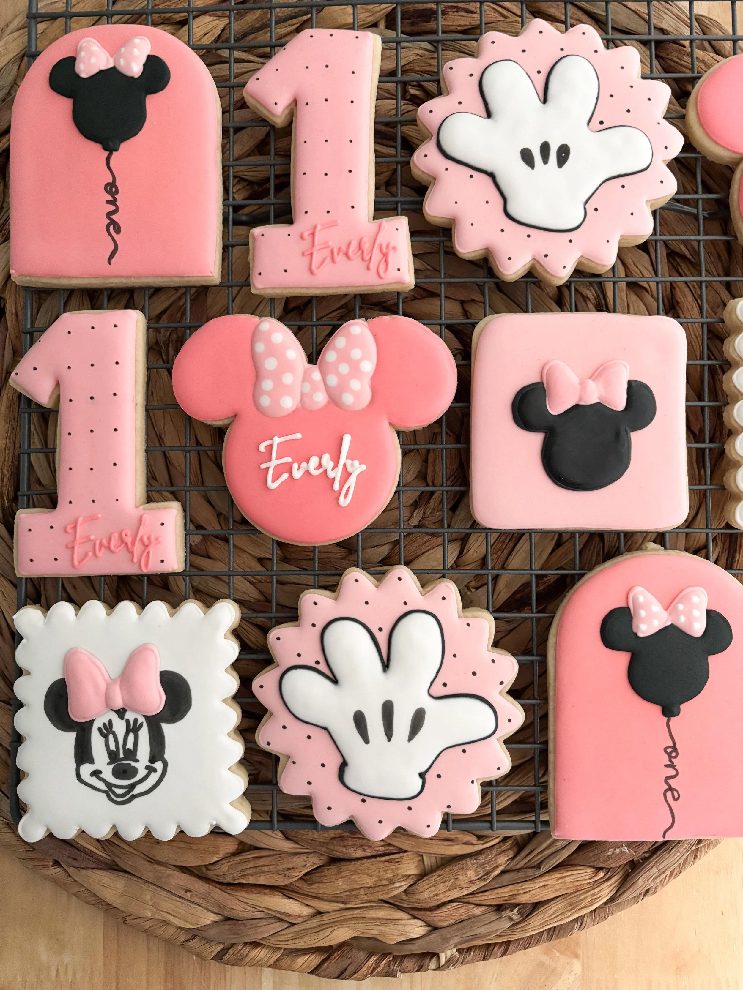 Decorated cookies with Minnie Mouse theme, including pink, white, and black cookies with mouse head silhouettes, bows, and the number one, placed on a wire cooling rack.