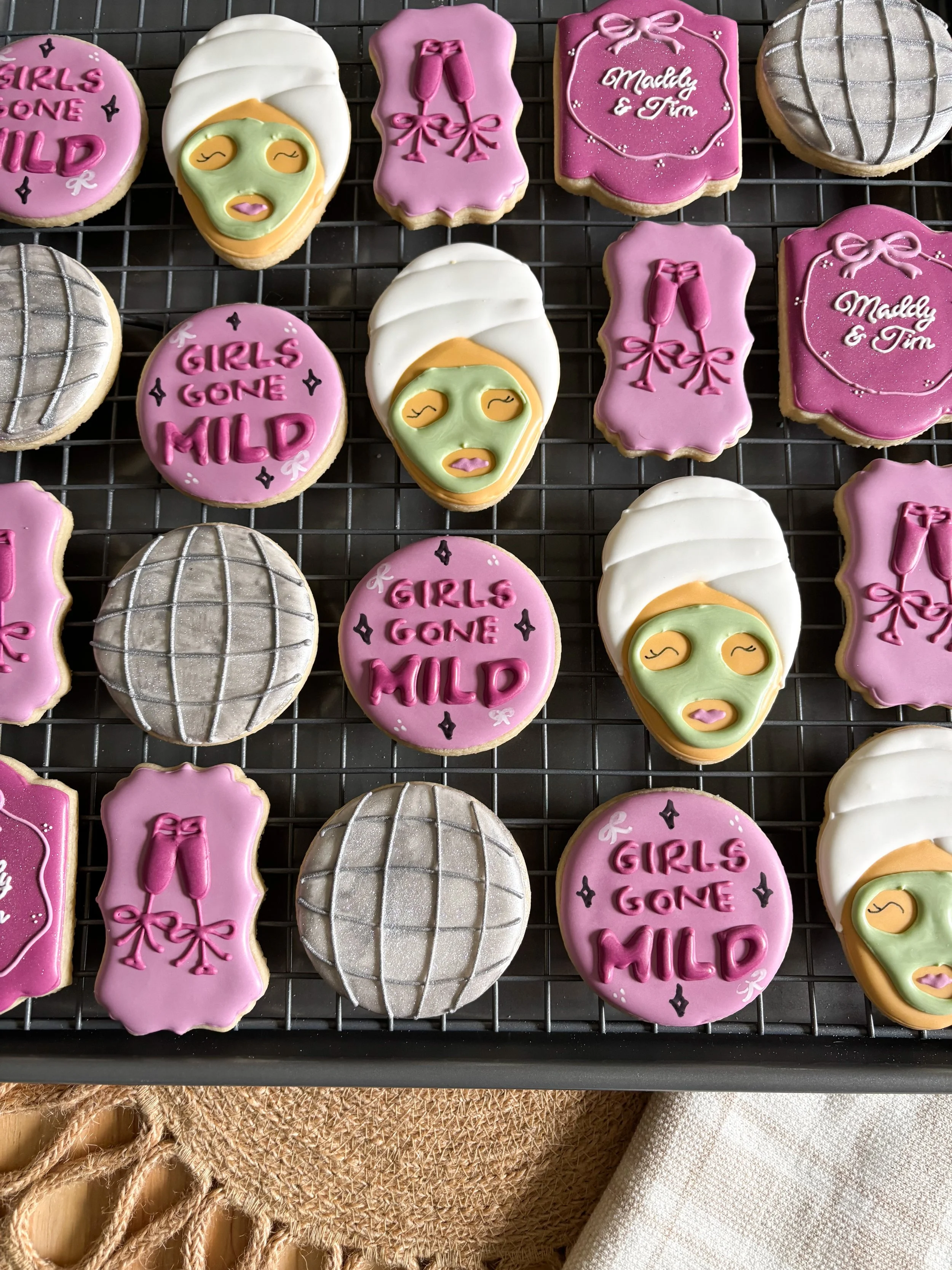 Decorated cookies on a cooling rack with messages 'Girls Gone MILD' and 'Madly & Tim', as well as lipstick and face mask designs in pink, white, and gray colors.
