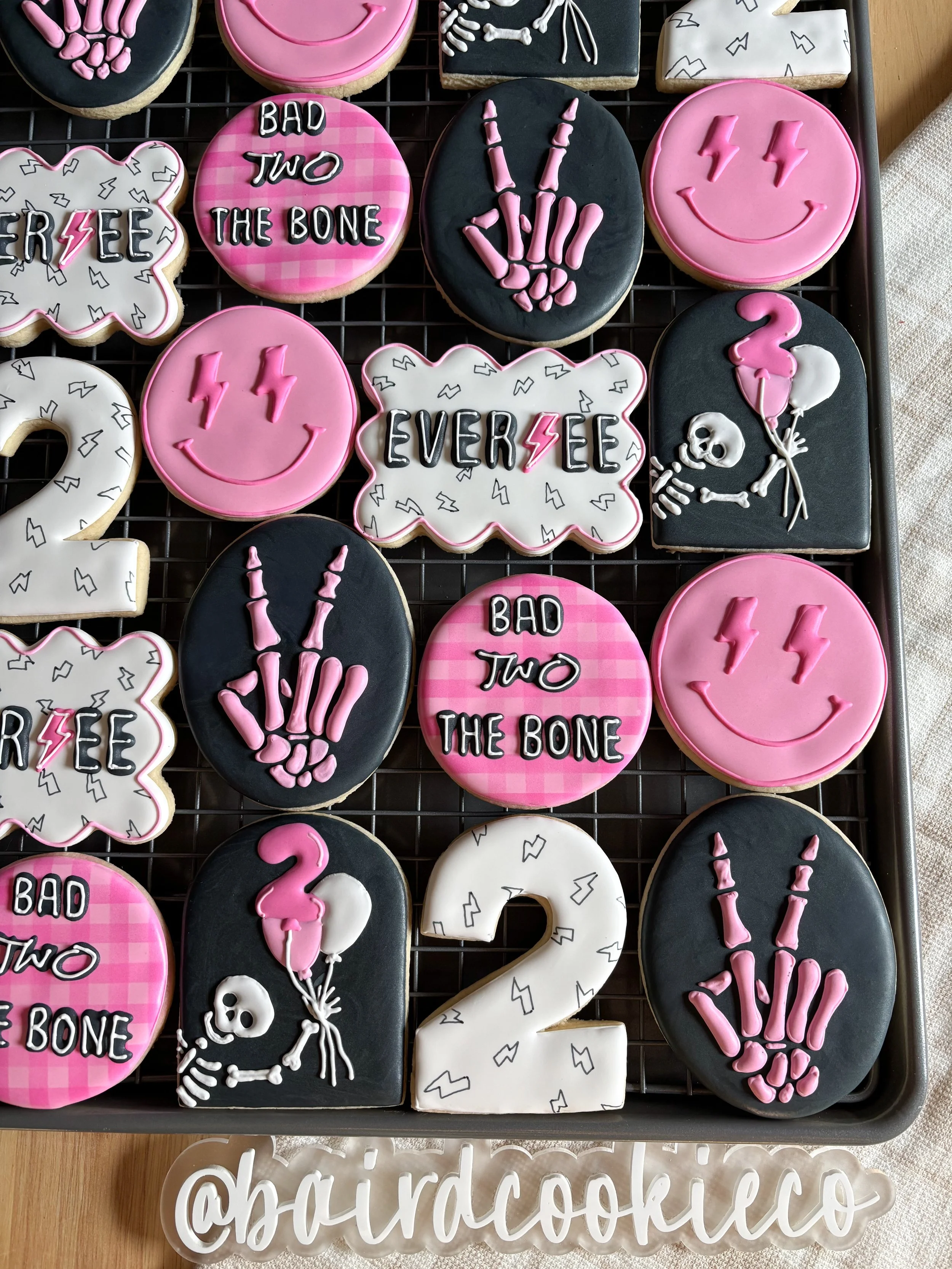 Decorated cookies with various designs including smiley faces, skeletons holding balloons, peace signs, phrases like "Bad Two The Bone," and numbers like "2" and "3," arranged on a cooling rack.