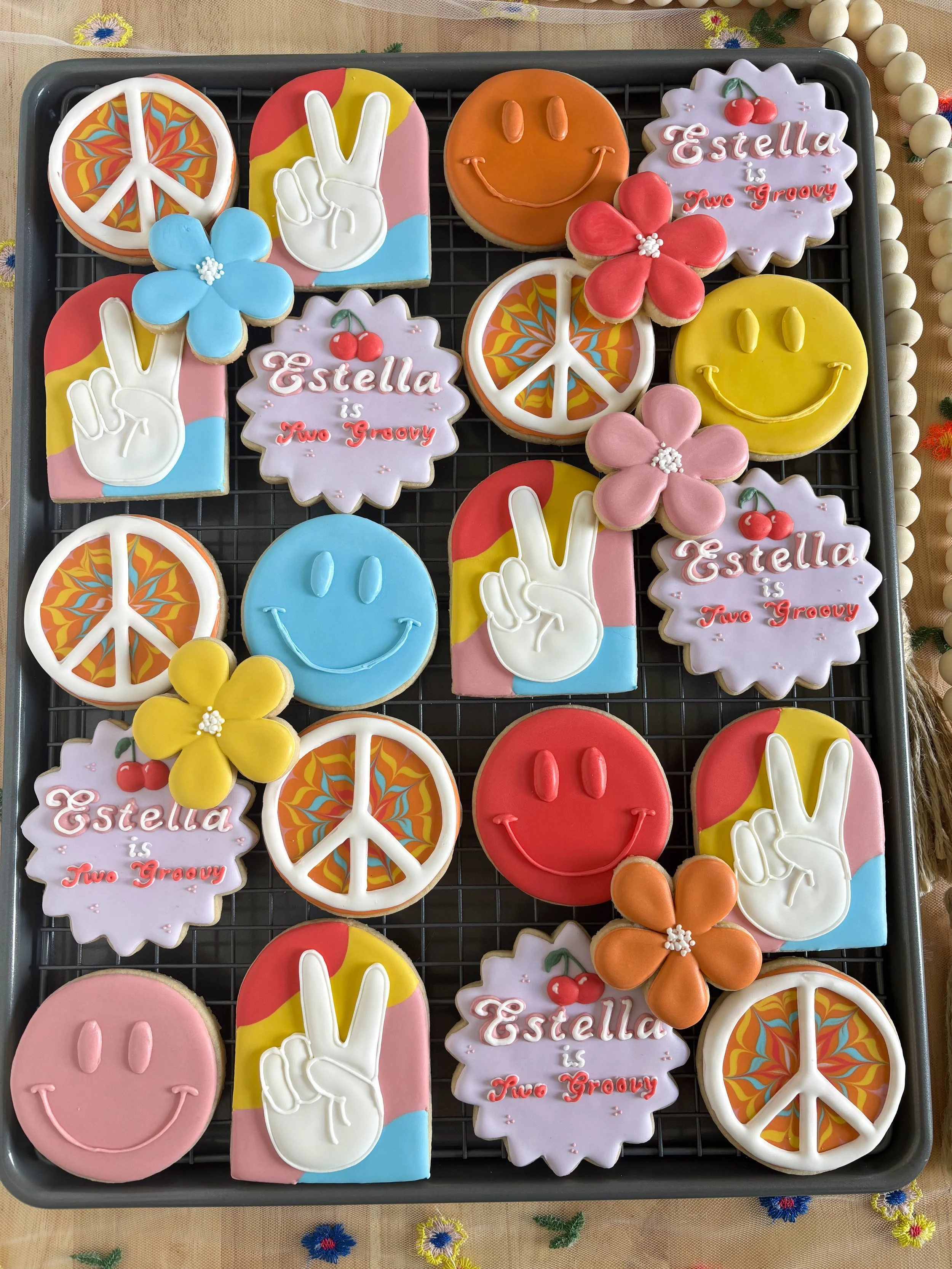 Decorated sugar cookies with peace signs, flowers, smiley faces, peace signs with colorful psychedelic patterns, peace signs with a star, and cookies with the text "Estella is Two Groovy" on purple backgrounds.