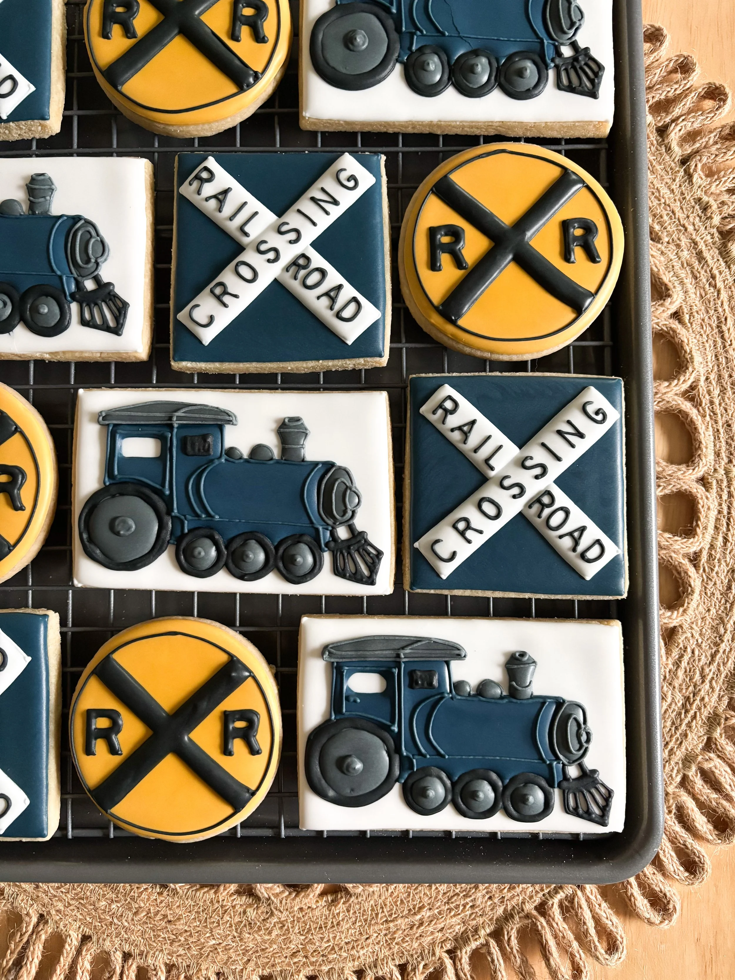 Cookies decorated with train, railroad crossing, and railroad signs in blue, yellow, black, gray, and white icing.