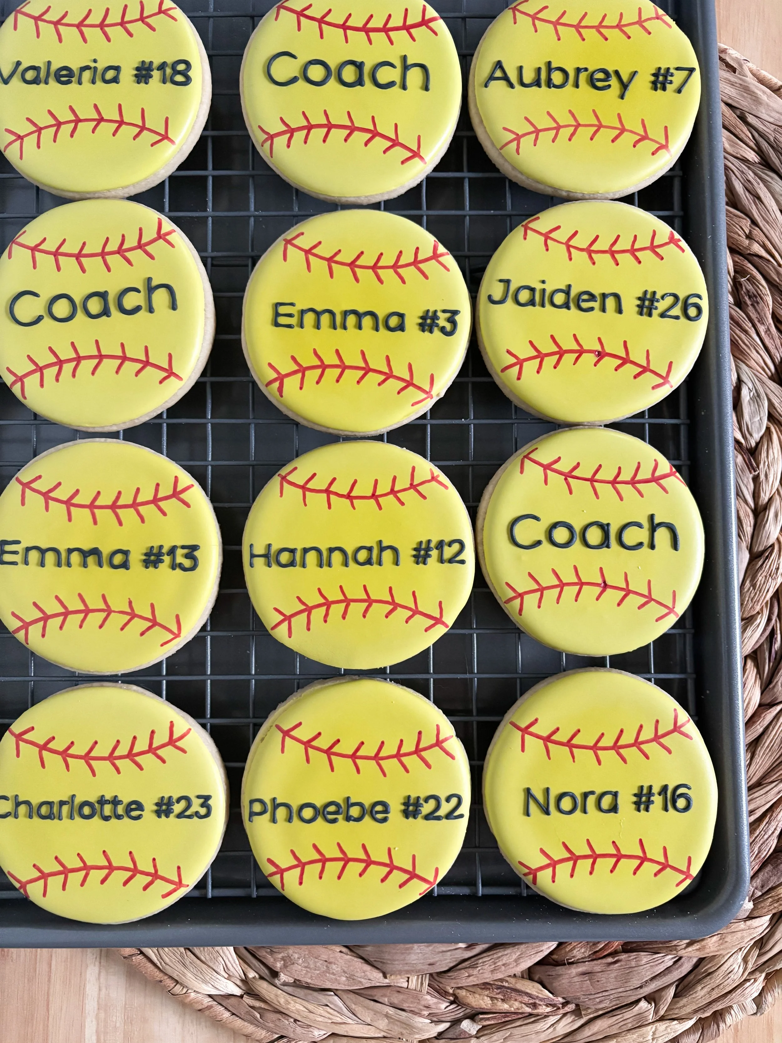 Decorative baseball-themed cookies with names and numbers written in black on yellow icing, arranged on a wire rack.