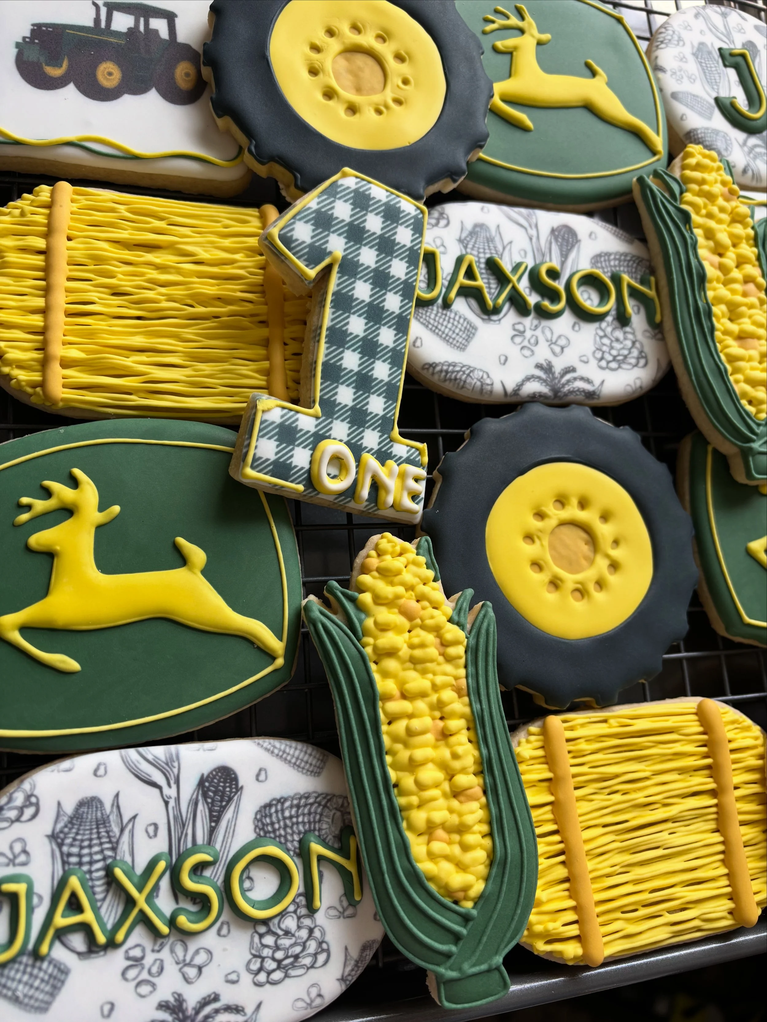 Decorated cookies for a first birthday celebration with a green and yellow color theme. The cookies feature images of a deer, a tractor, a tire, a corn cob, and a plaid number 1 with the name 'Jaxson' and the word 'One'.