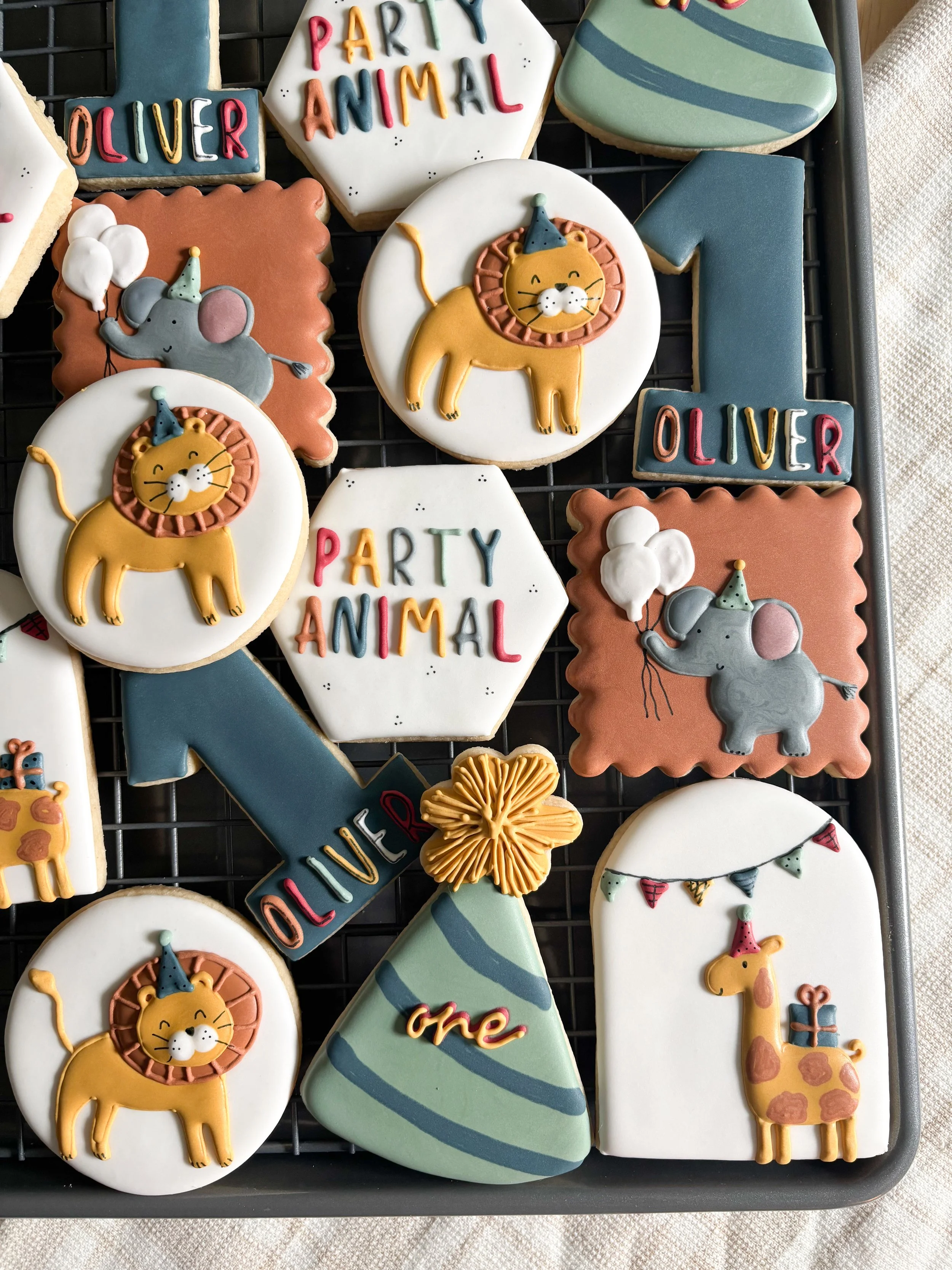 Decorated cookies for a first birthday celebration with a circus animal theme. The cookies feature lions, elephants with balloons, giraffes with presents, party hats, and banners with the name Oliver and the phrase 'Party Animal.'