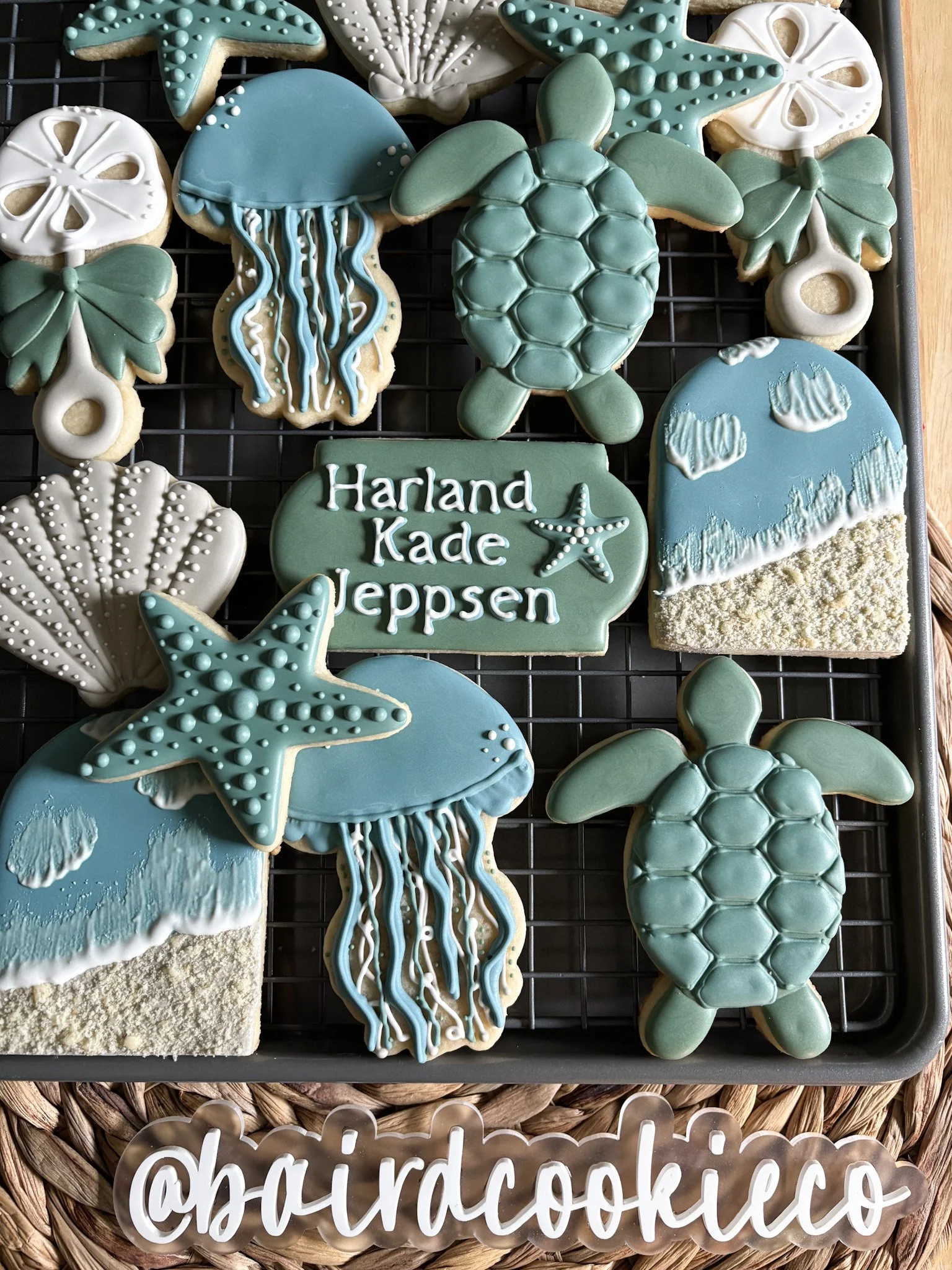 Assorted beach-themed decorated cookies on a cooling rack, including turtles, jellyfish, starfish, seashells, and a cloud scene, with a personalized text cookie that says "Harland Kade Jeppsen".
