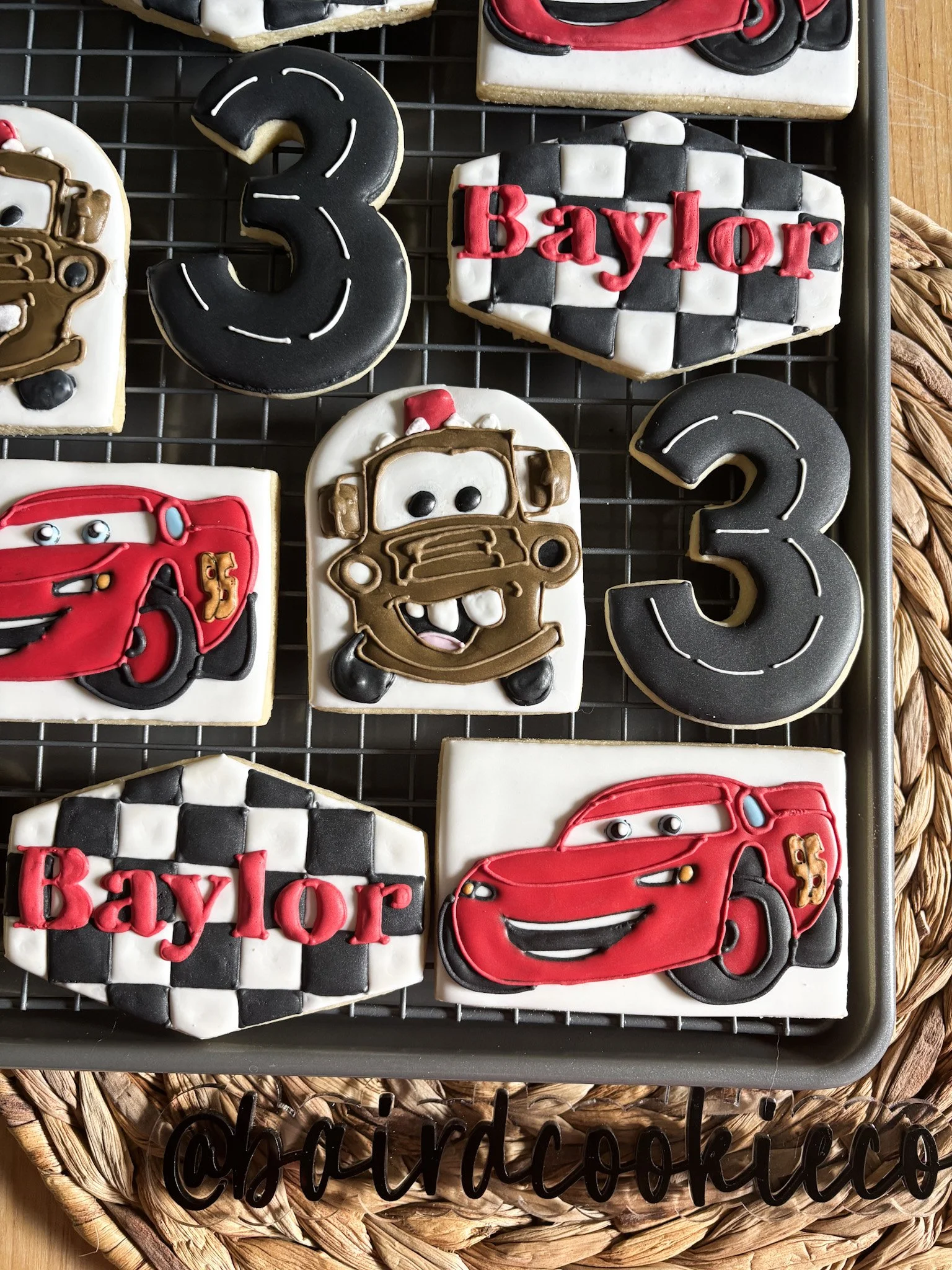 Decorated cookies featuring characters from Cars, a checkered pattern with the word 'Baylor', and the number 3, arranged on a cooling rack.