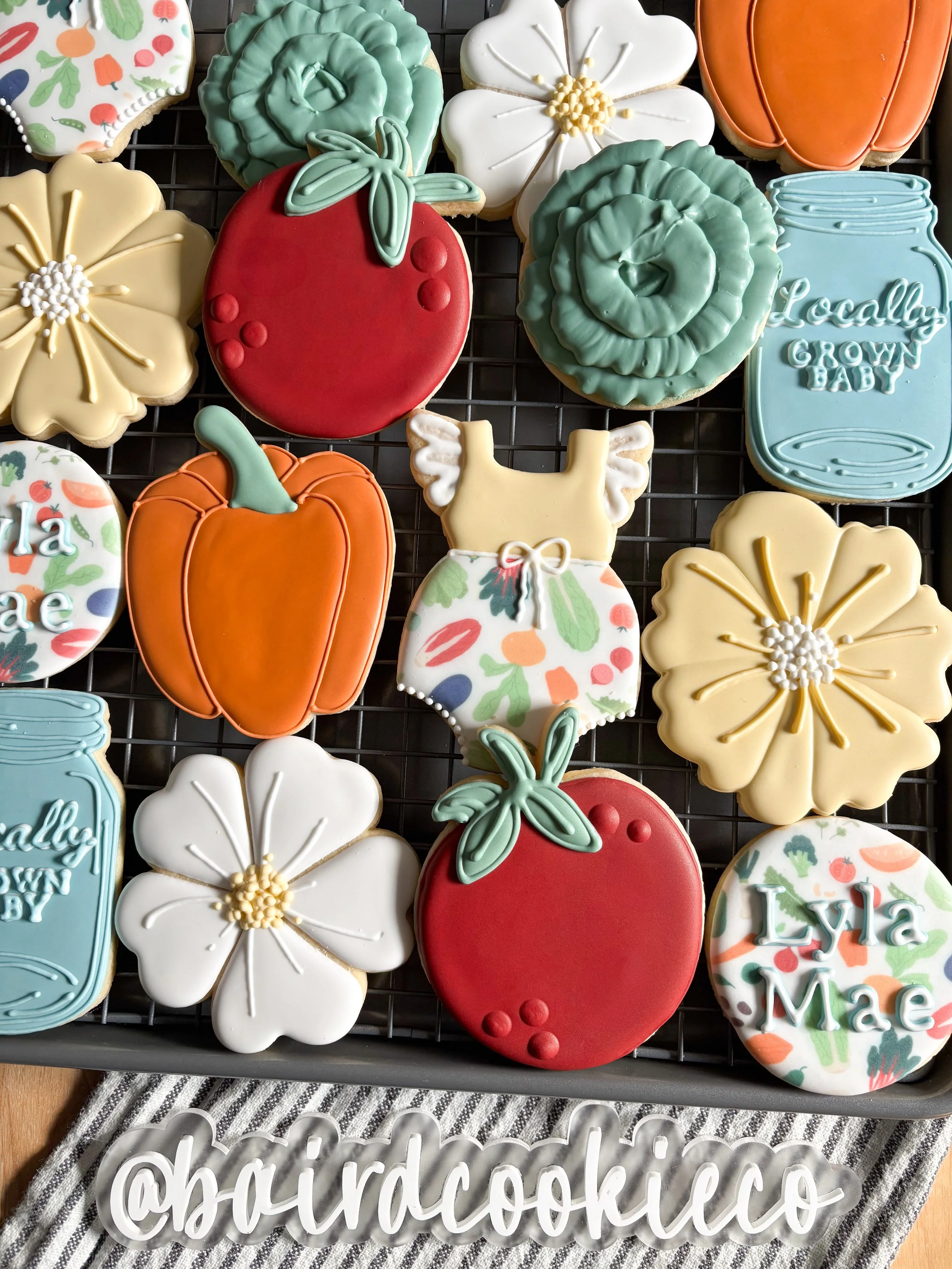 Decorated cookies shaped as fruits, flowers, pumpkins, and a dress, with colorful icing, displayed on a cooling rack.