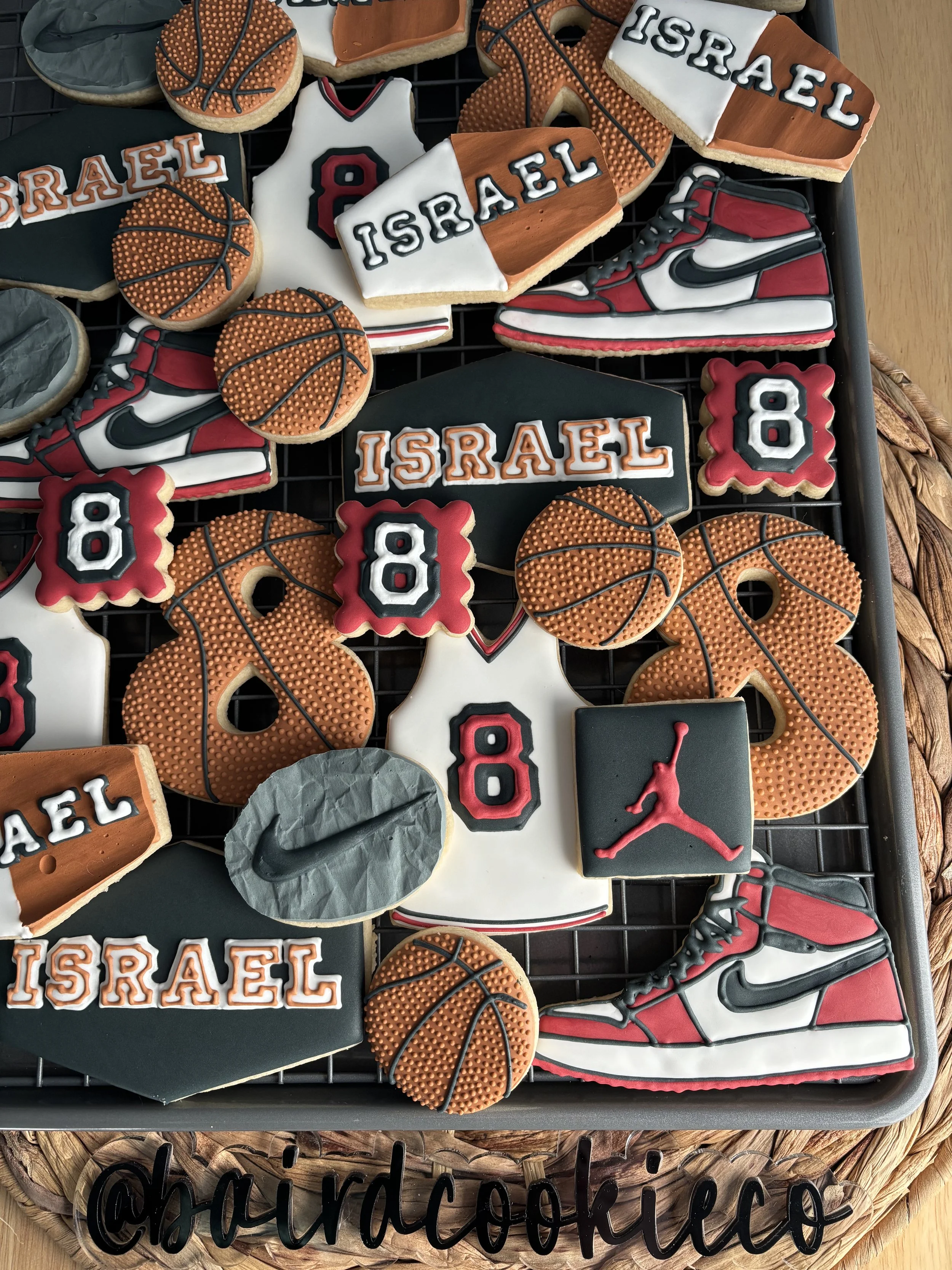 Assorted basketball-themed and sports-themed decorated cookies, including cookies with basketball designs, Nike swoosh logos, the word 'Israel,' and the number 8, arranged on a cooling rack.