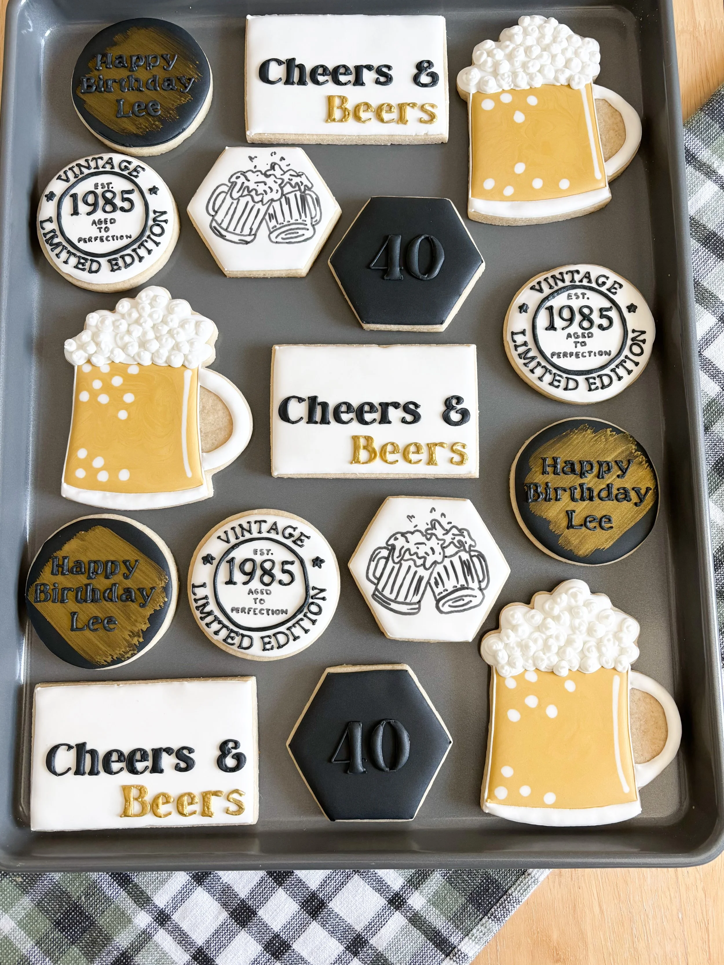 A tray of decorated birthday cookies with beer and celebration-themed designs, including messages like 'Happy Birthday Lee' and 'Cheers & Beers', and a cookie with '40' on it, on a checkered tablecloth.