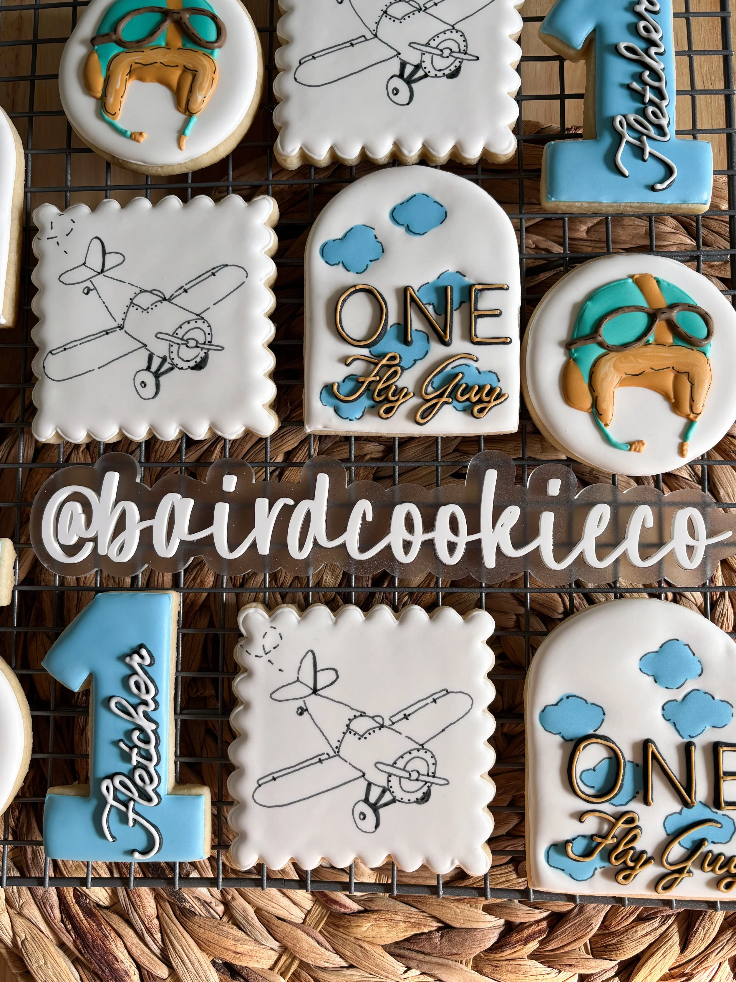 Decorative cookies with airplane and sky themes, featuring messages like "ONE Fly Guy" and Instagram handle '@boardcookie', arranged on a wire cooling rack.