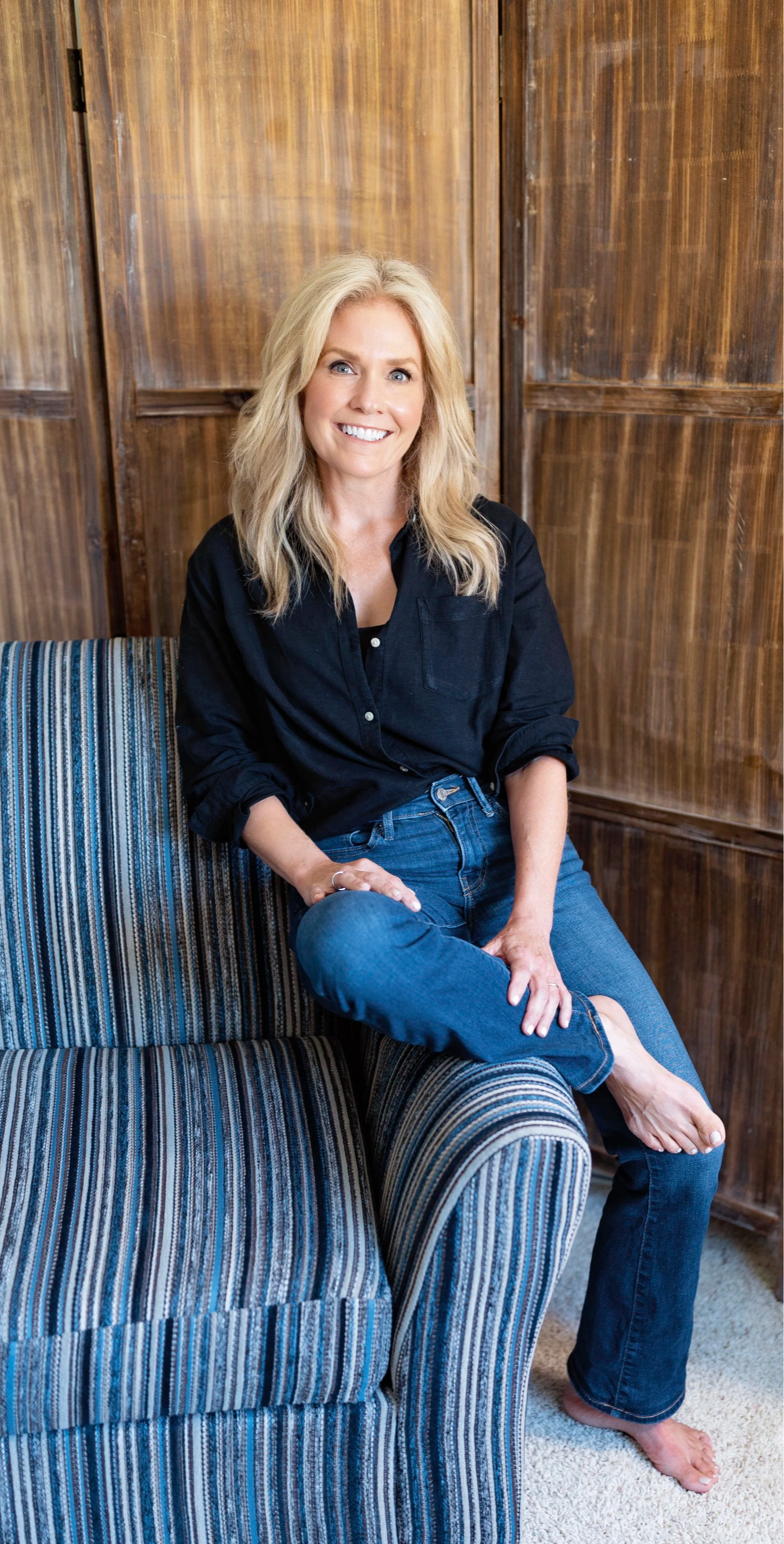 A woman with blonde hair, wearing a black button-up shirt and blue jeans, sitting on a striped couch in front of a wooden wall and smiling.