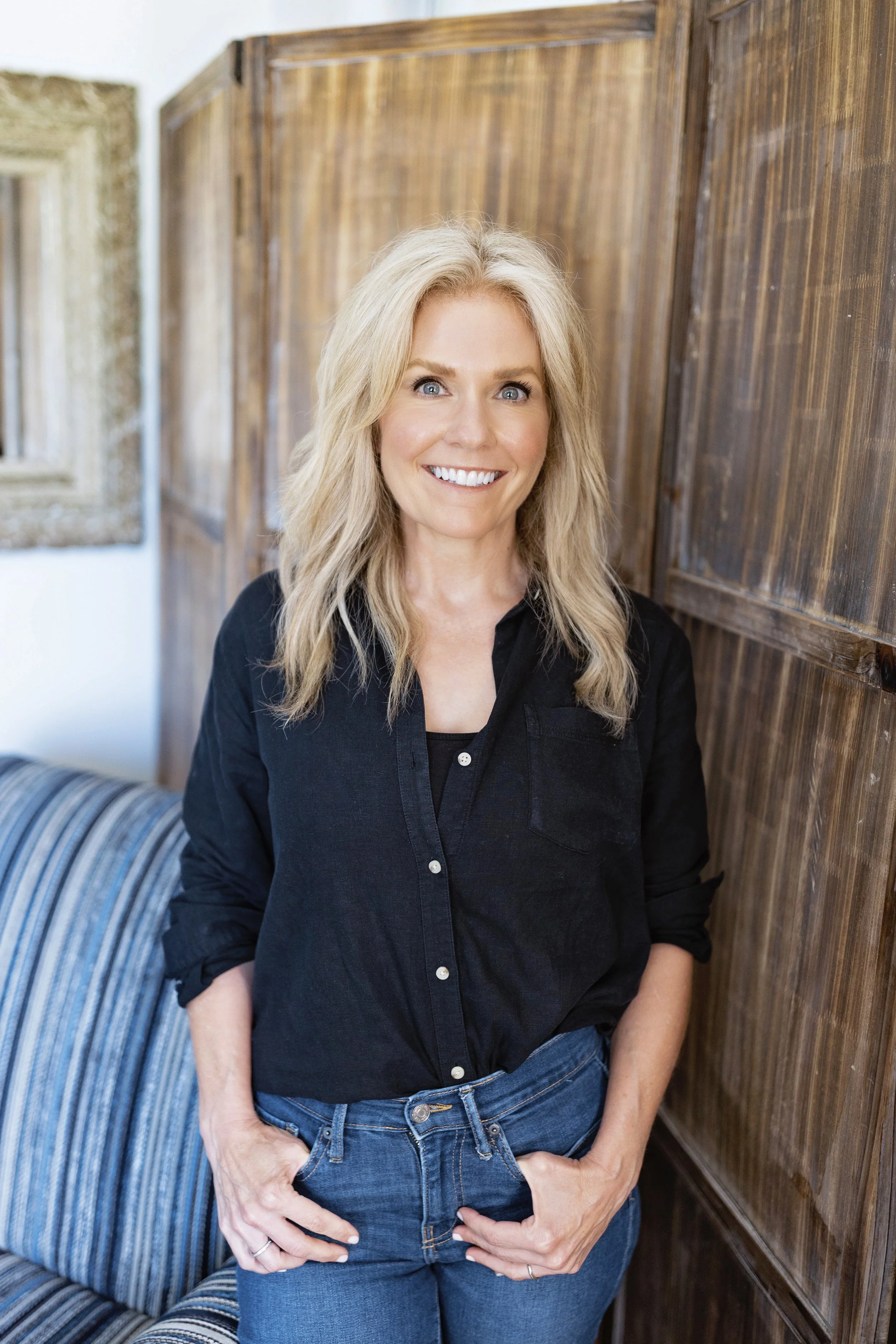 A blonde woman smiling, wearing a black shirt and blue jeans, standing indoors against a wooden paneled wall.