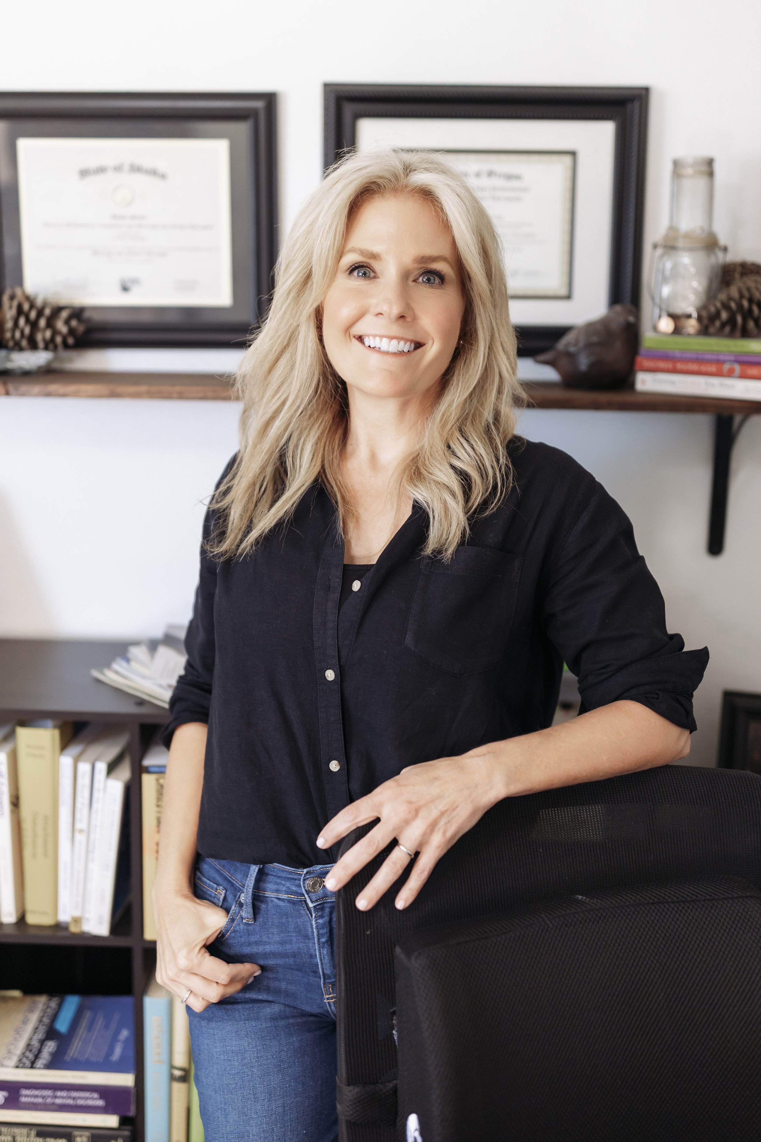 A woman with blonde hair wearing a black shirt, standing in an office, smiling, with framed certificates on the wall behind her.