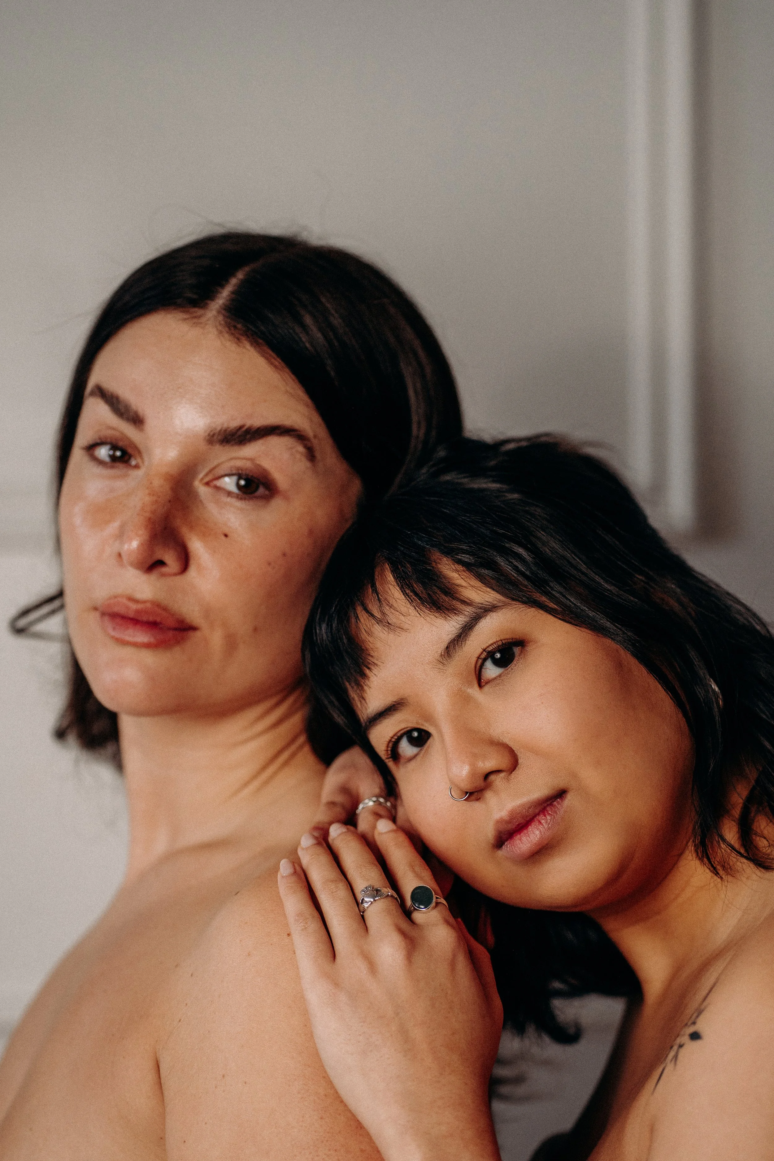 Close-up photo of two women, one with light skin and dark brown hair, and the other with Asian features and black hair, embracing affectionately. The woman with dark hair has a septum piercing and multiple rings on her fingers.