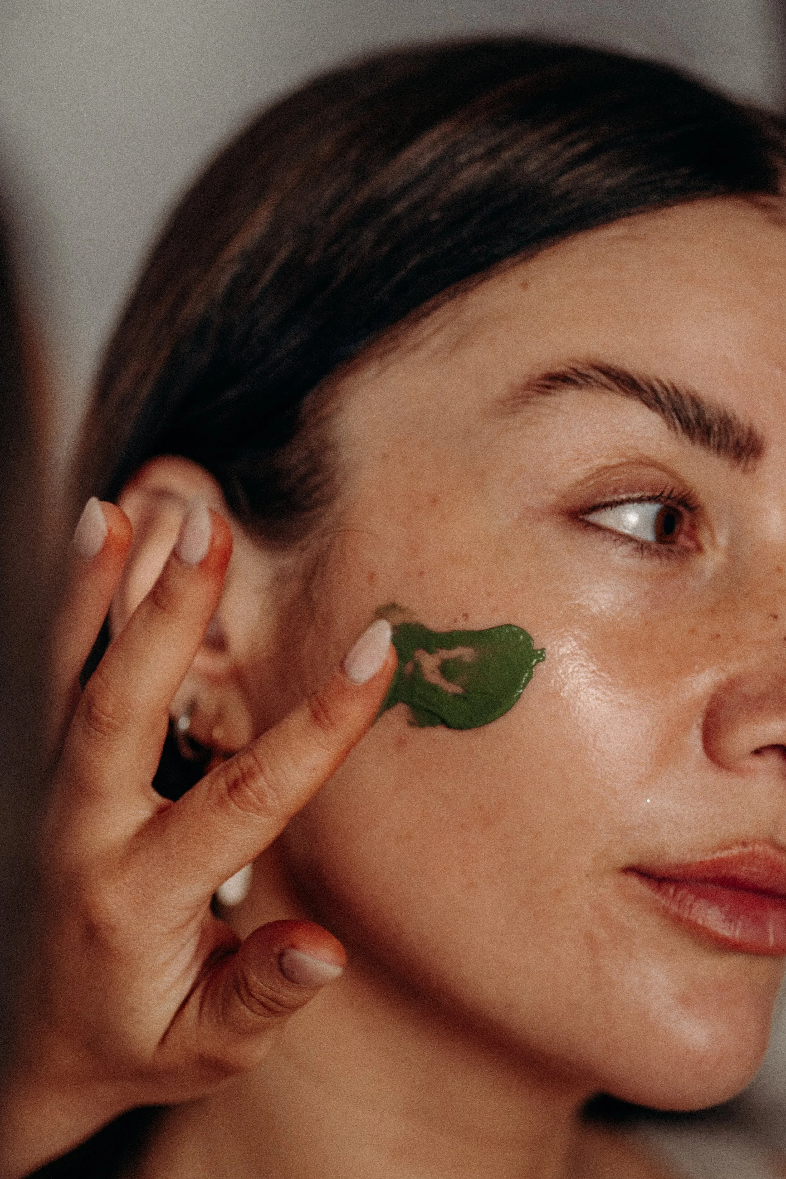 Close-up of a woman with dark brown hair, using her fingers to apply a green facial mask on her cheek.