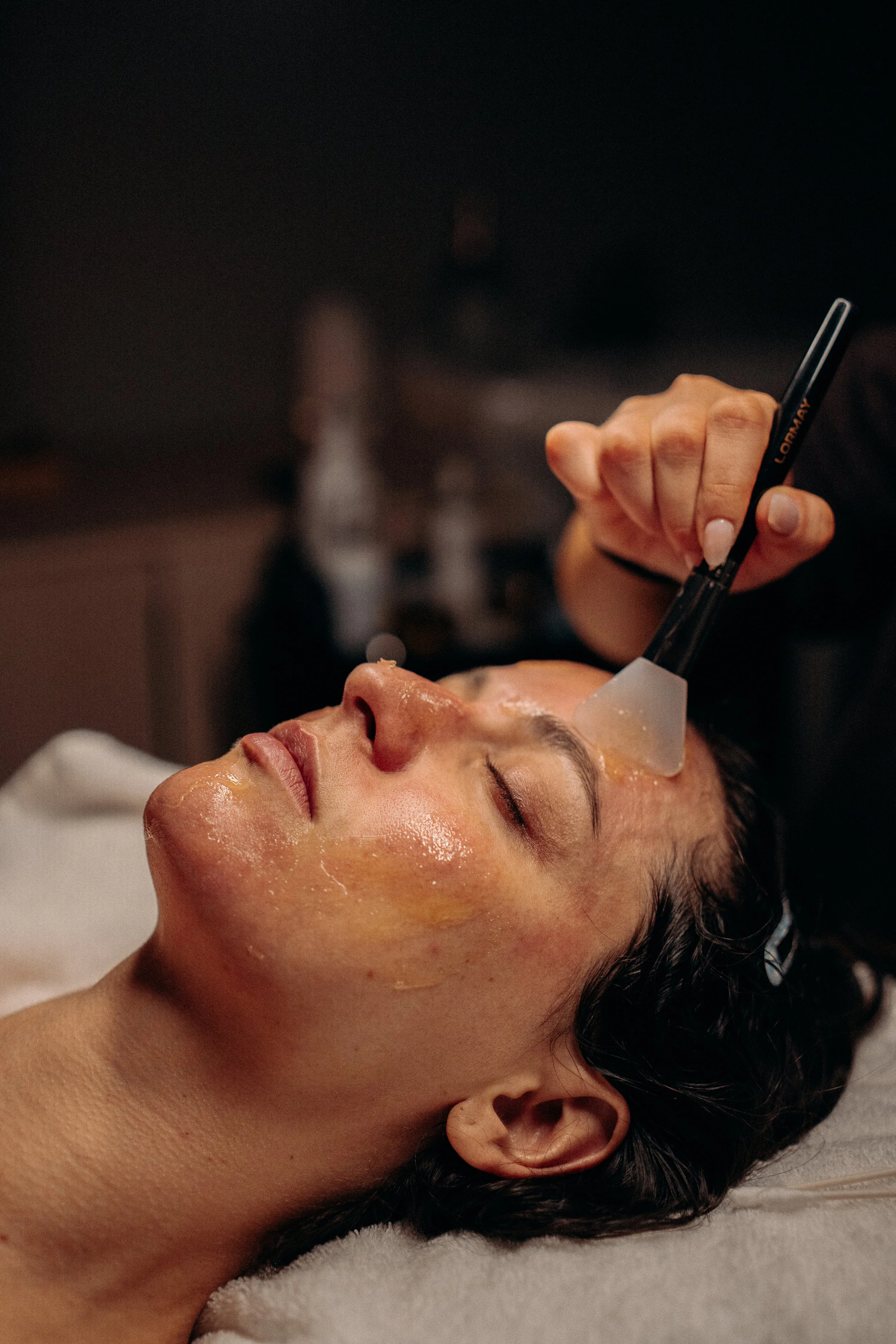 A woman receiving facial treatment with a black pen and a plastic mask on her face, lying down with her eyes closed.