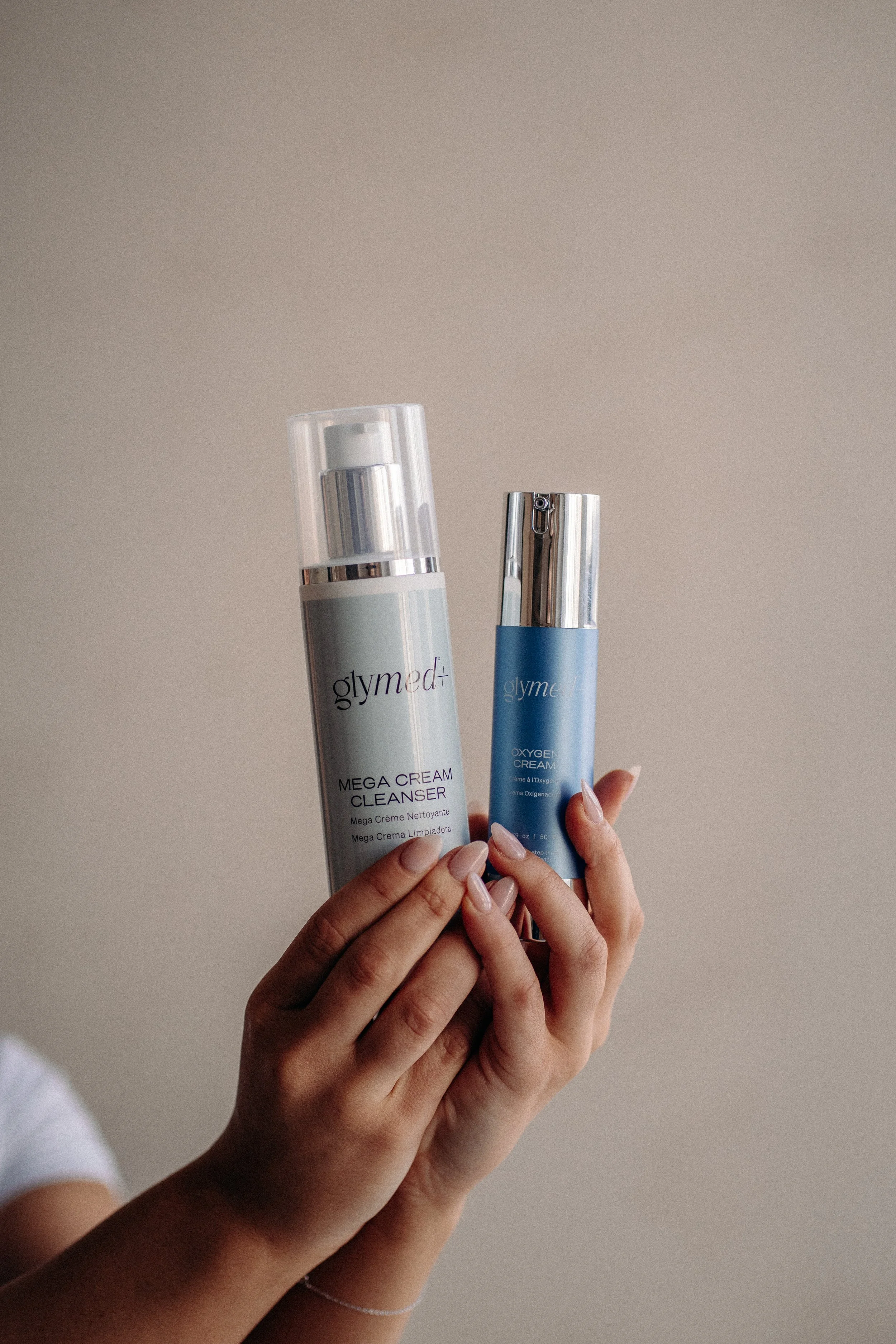 A person with light skin holds two skincare products; a white bottle labeled 'glymed+', 'Mega Cream Cleanser', and a blue bottle labeled 'glymed+', 'Oxygen Cream' against a plain beige background.