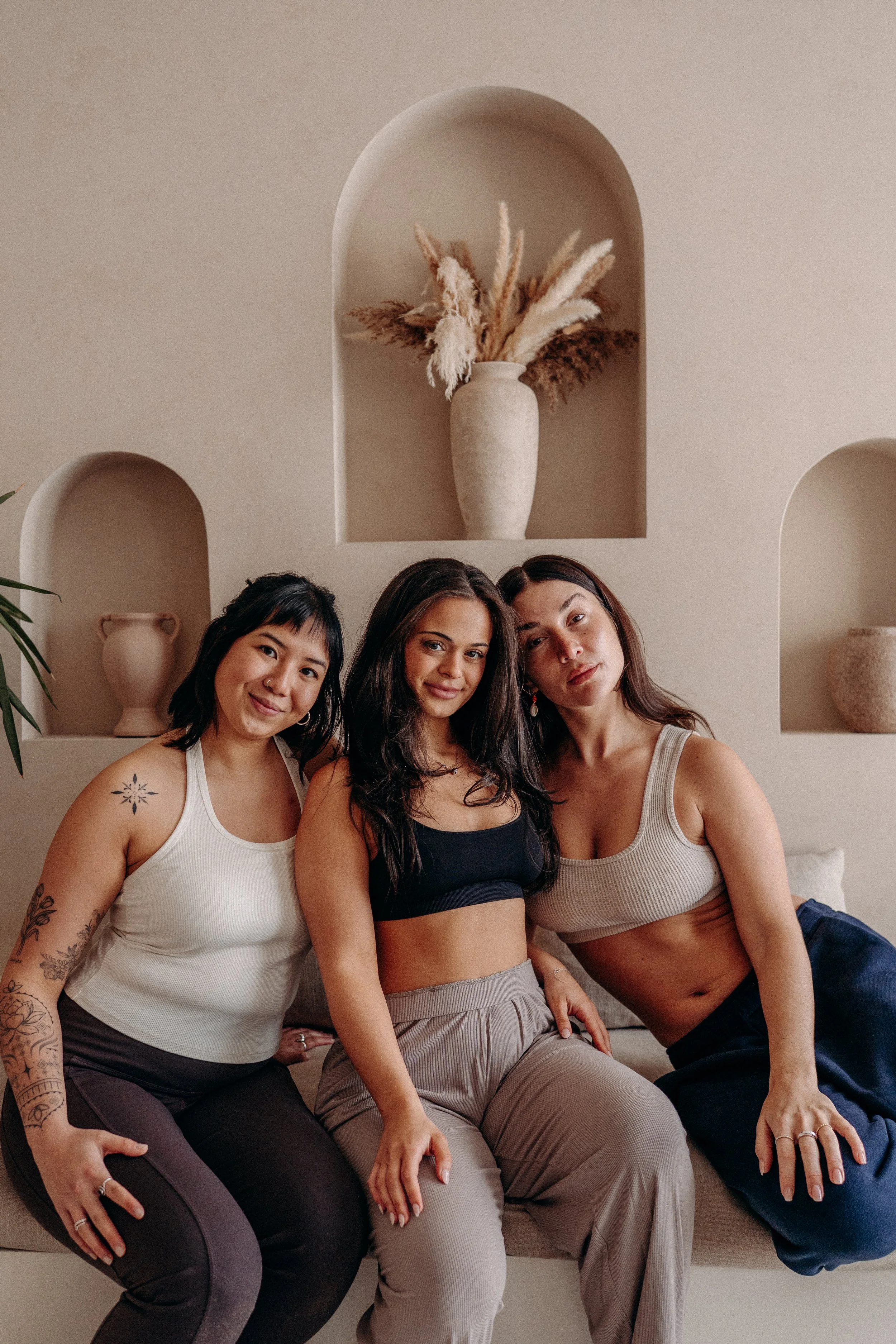 Three women sitting closely together on a beige bench in a minimalistic room with arched wall niches. There is a beige vase with dried pampas grass in the center niche. The women are in relaxed poses, wearing casual tank tops and pants, and smiling softly.