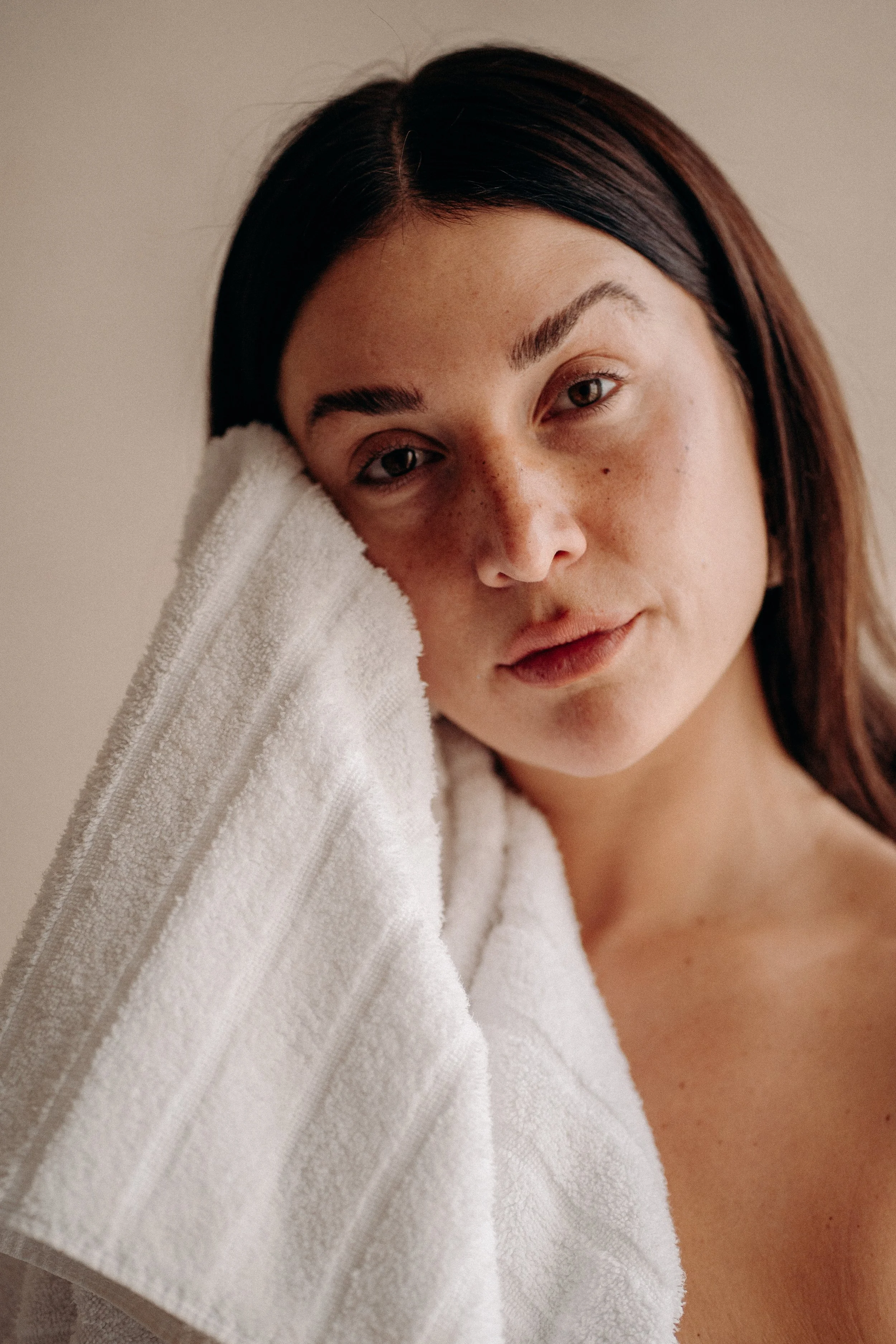 Close-up of a young woman with dark hair, resting her face on her hand, with a white towel draped over her shoulder, against a neutral background.