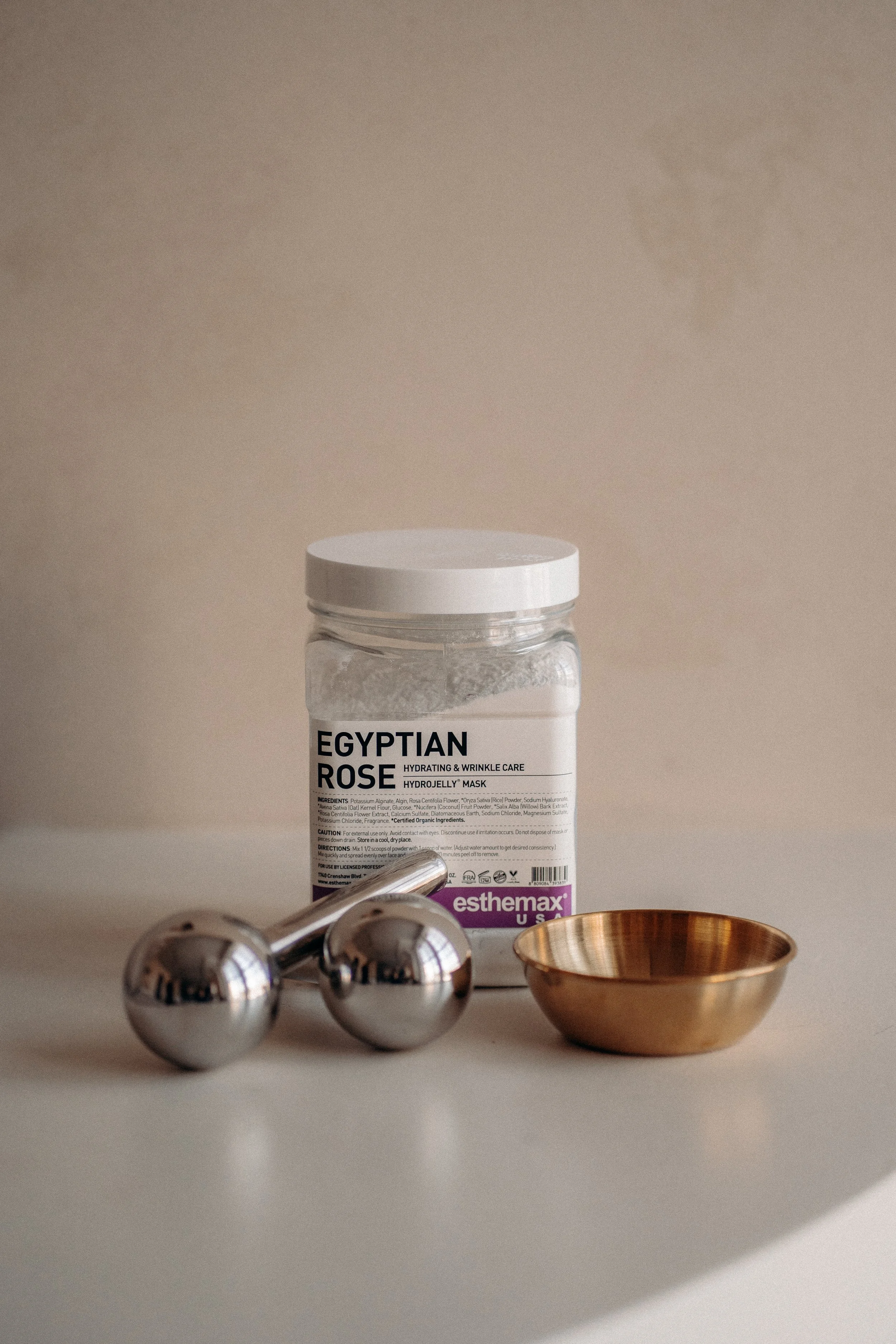 A white bottle labeled 'Egyptian Rose Hydrating & Wrinkle Care Hydrojelly Mask' on a beige background. In front of it are three shiny metallic massage balls, and to the right is a small, round, gold-colored bowl.
