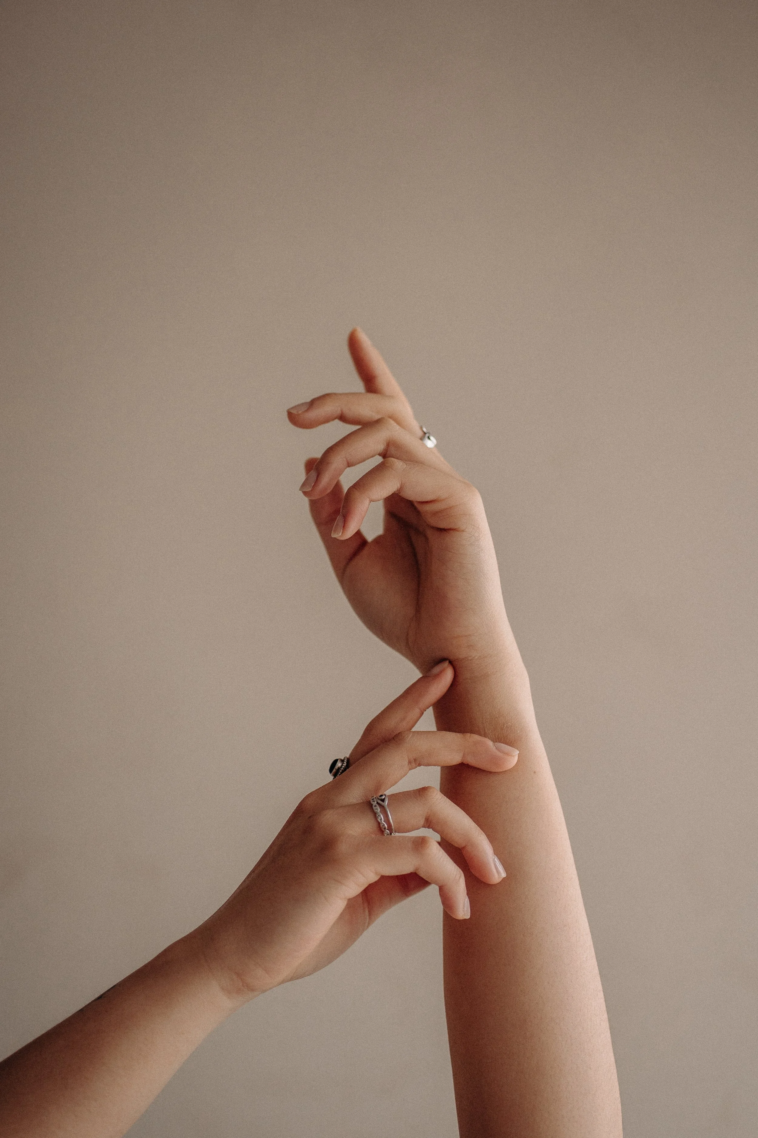 Multiple hands with rings touch near the wrist area against a beige background.