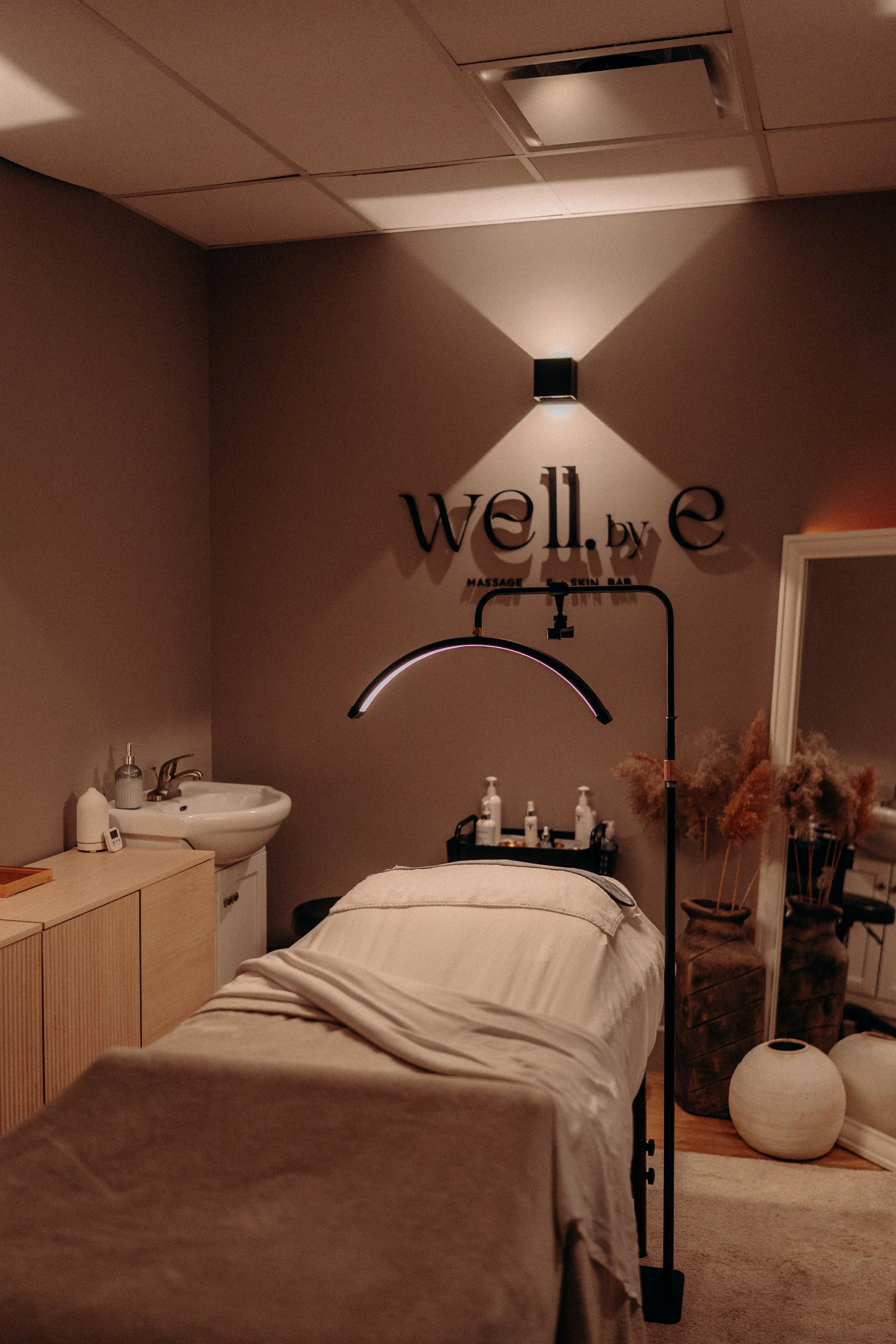 A massage room with a massage table covered in white sheets, a small wooden cabinet, a sink, and decorative vases with dried plants. There is a wall-mounted light fixture and a sign that reads 'well.by.c' with smaller text underneath that says 'massage,' 'facial,' 'skin bar,' and 'body treatments.'