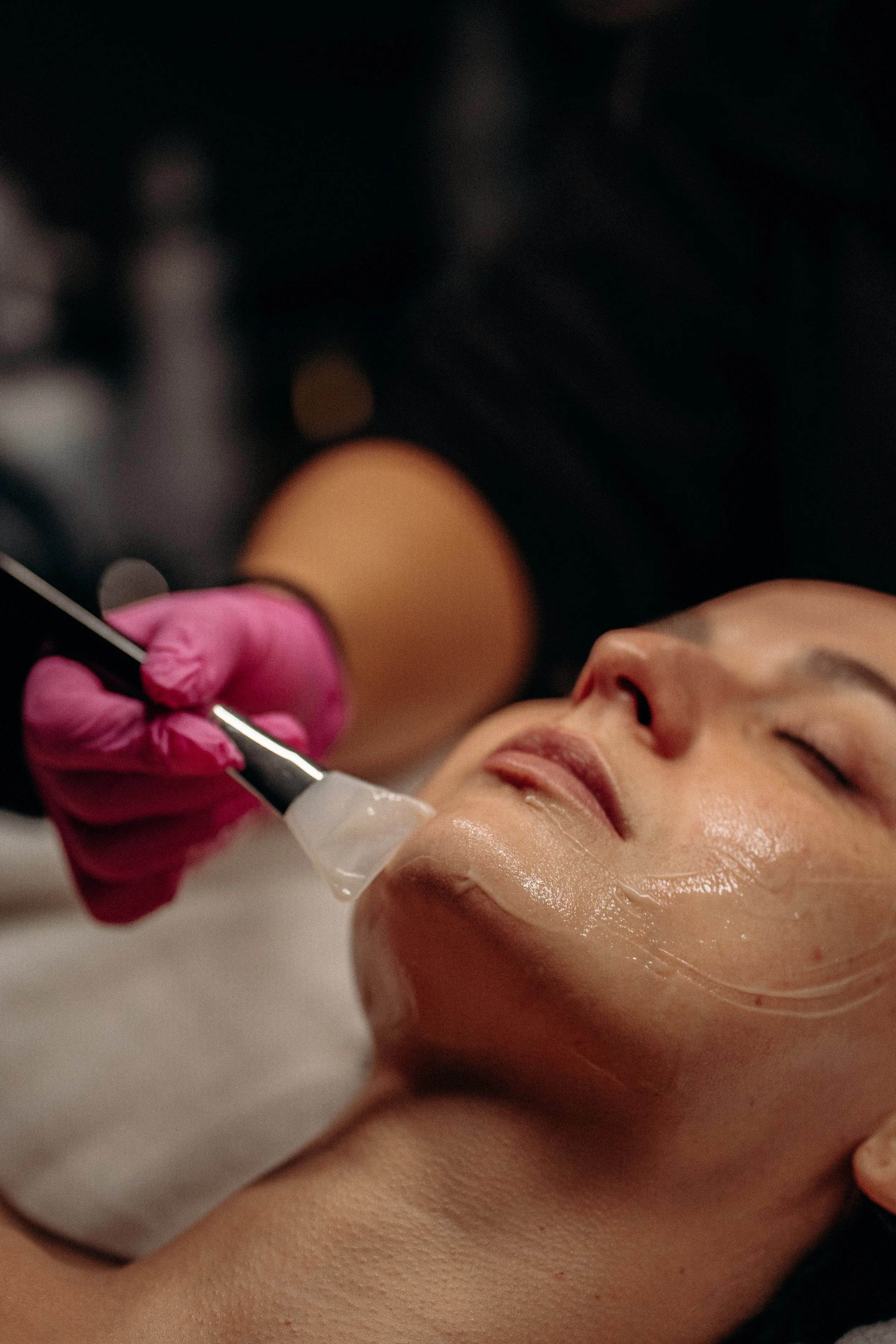 Close-up of a woman receiving a soothing facial mask on her face, lying down with closed eyes and a gloved hand administering the mask.