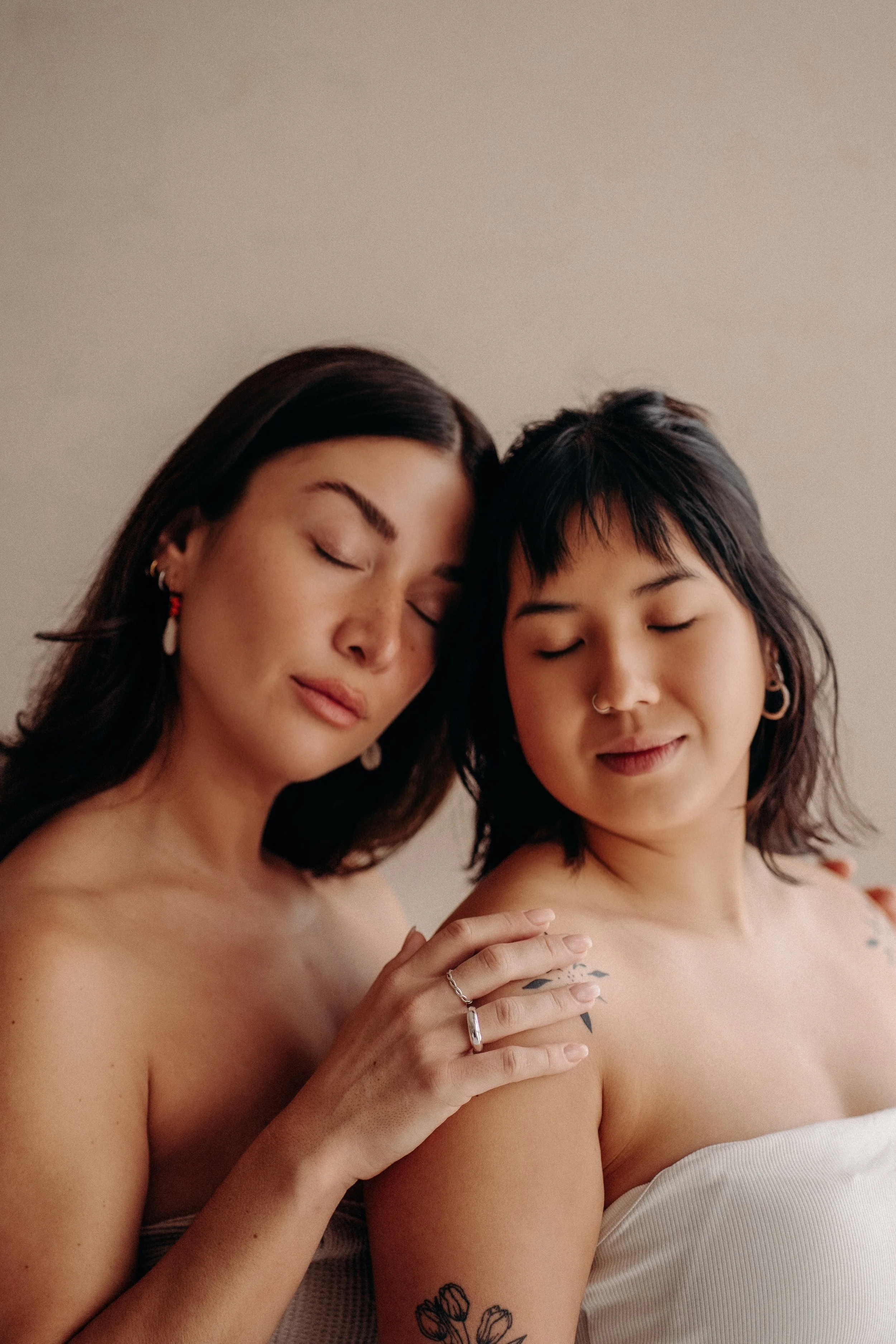 Two women with short dark hair and tattoos on their arms close together, both with eyes closed and serene expressions, against a plain light background.