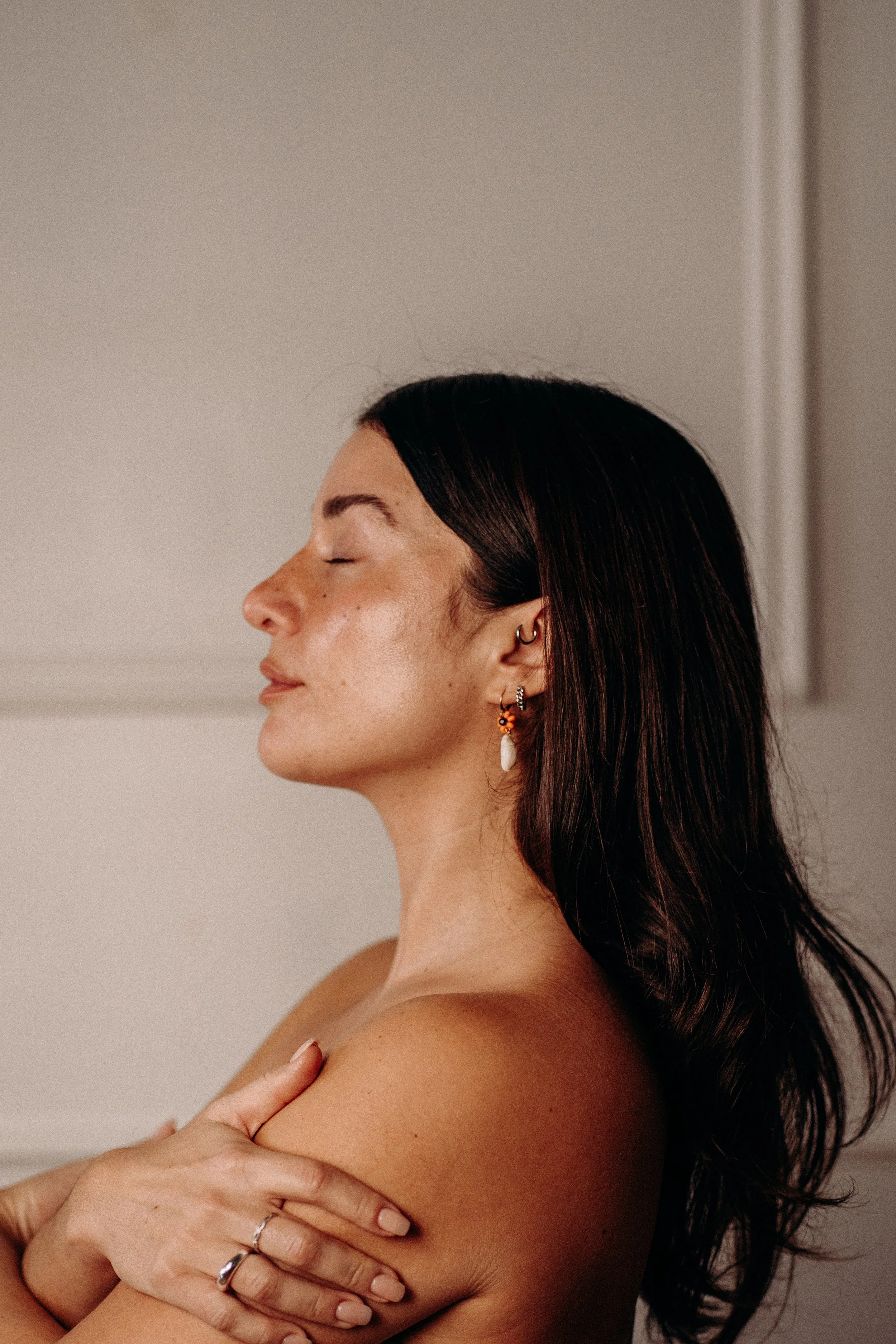 A woman with long dark hair and multiple earrings, including a hoop and a floral design, is hugging herself with eyes closed against a neutral background.