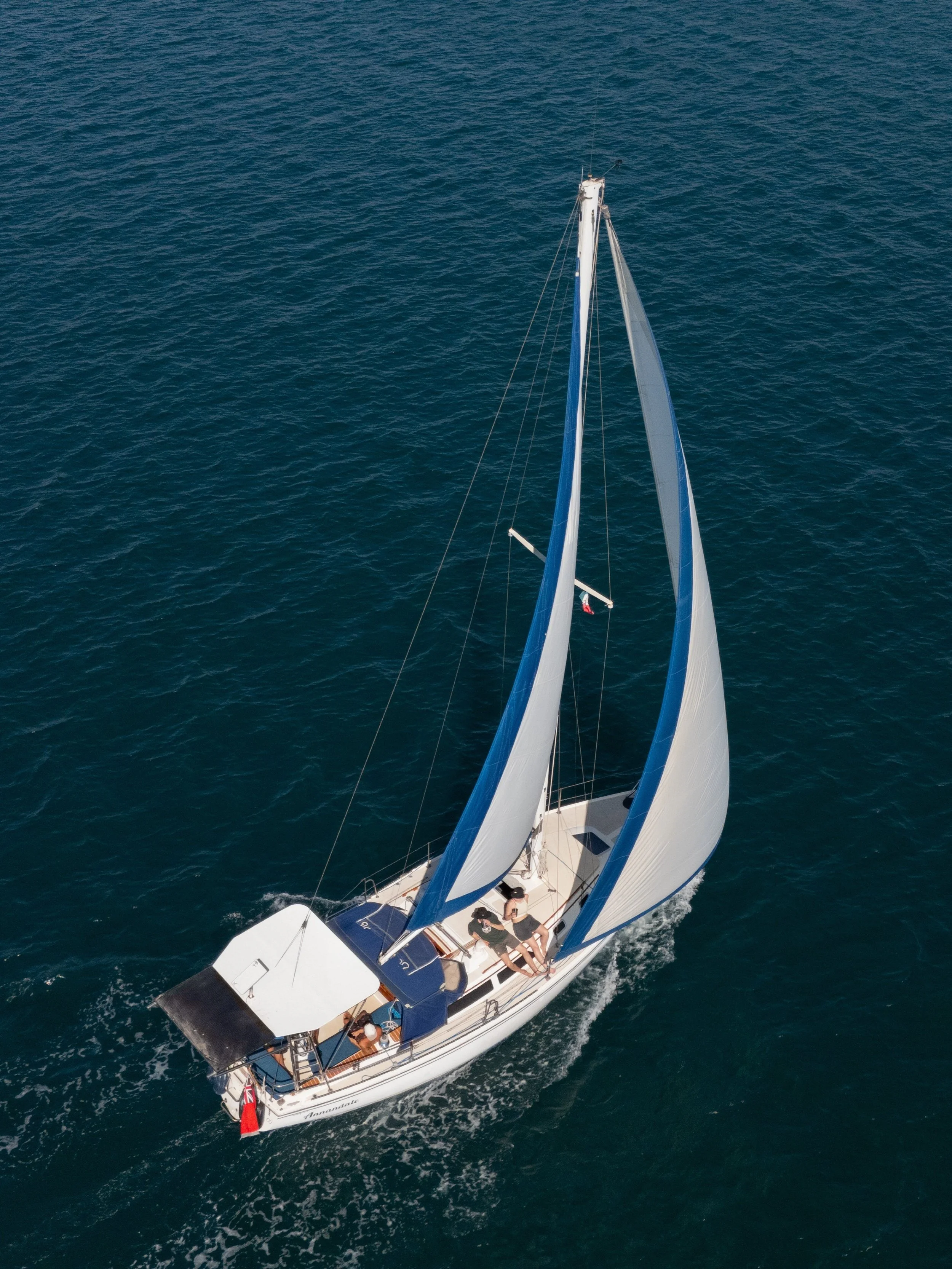 An aerial view of a sailboat with blue and white sails on the ocean, carrying four people on deck.