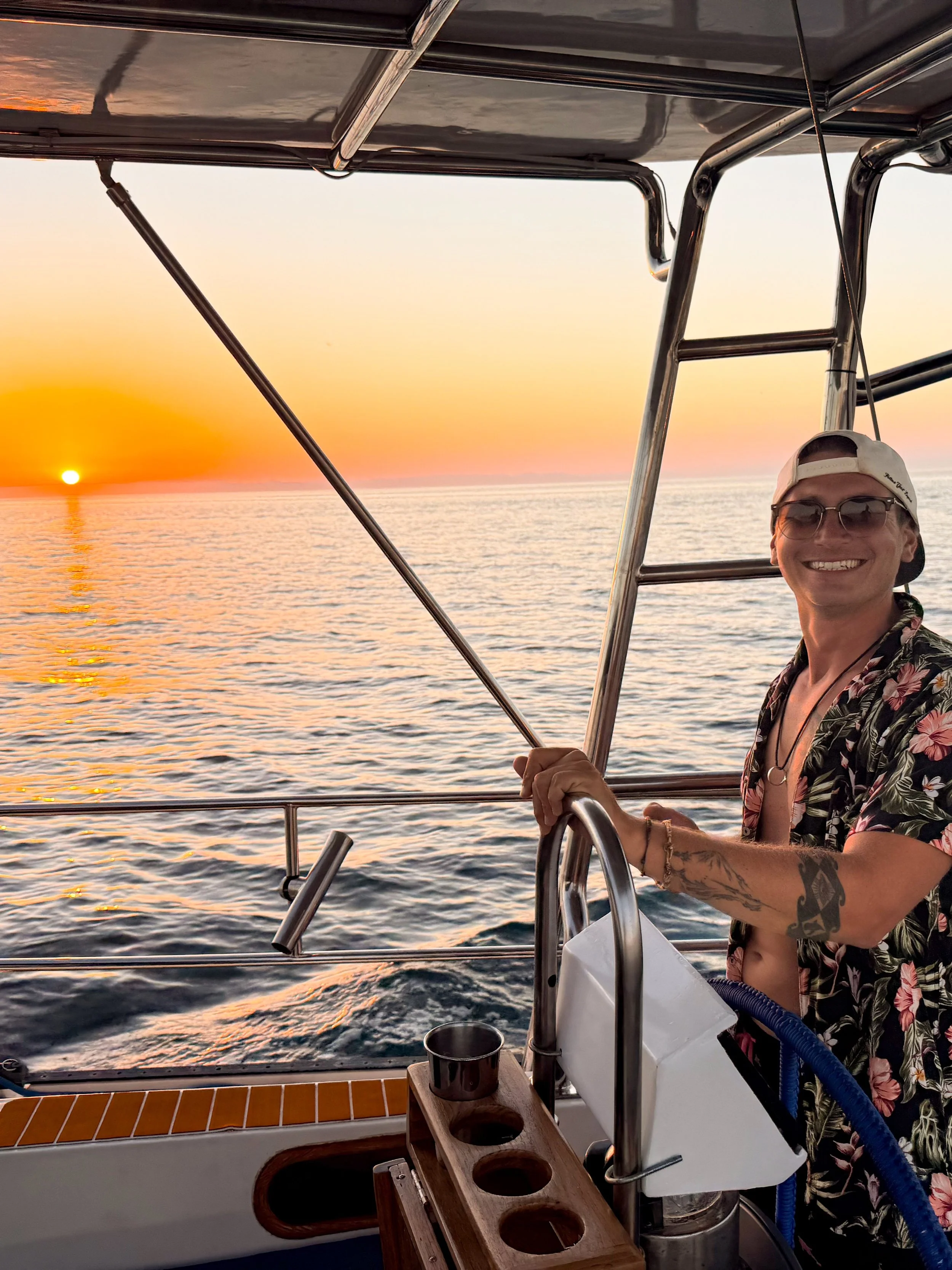 A person smiling on a boat during sunset with the ocean in the background. The person is wearing sunglasses, a cap, and a floral shirt, standing near boat equipment.