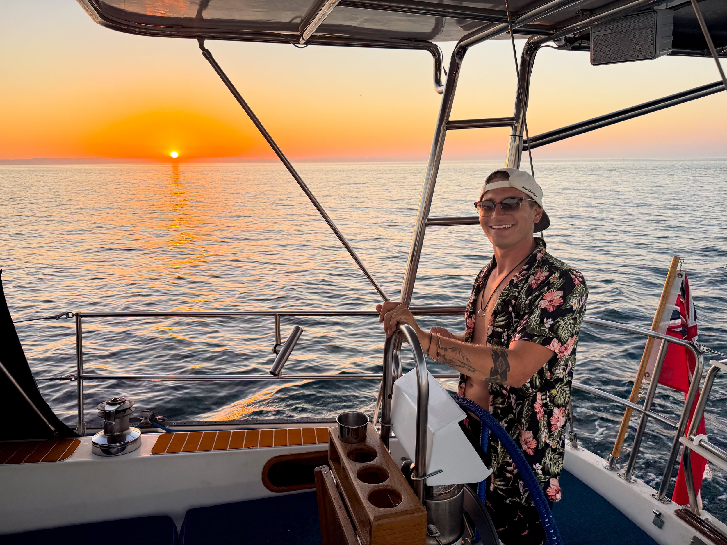 A smiling man wearing sunglasses, a white cap worn backwards, and a floral shirt, steering a boat on the water during sunset with an orange sky and the sun near the horizon.