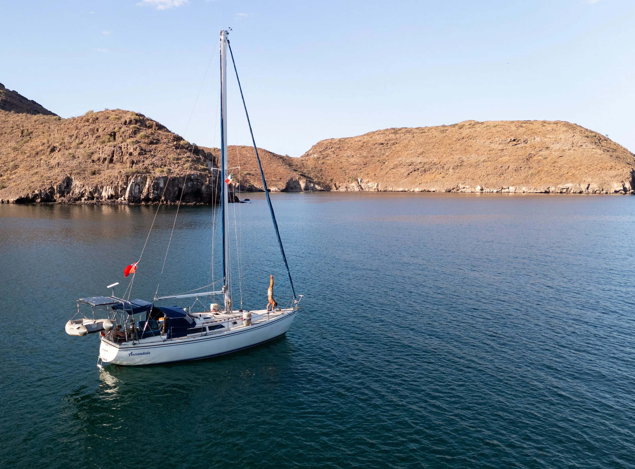 A sailboat named Annandale floating on calm water near rocky hills under clear blue sky.