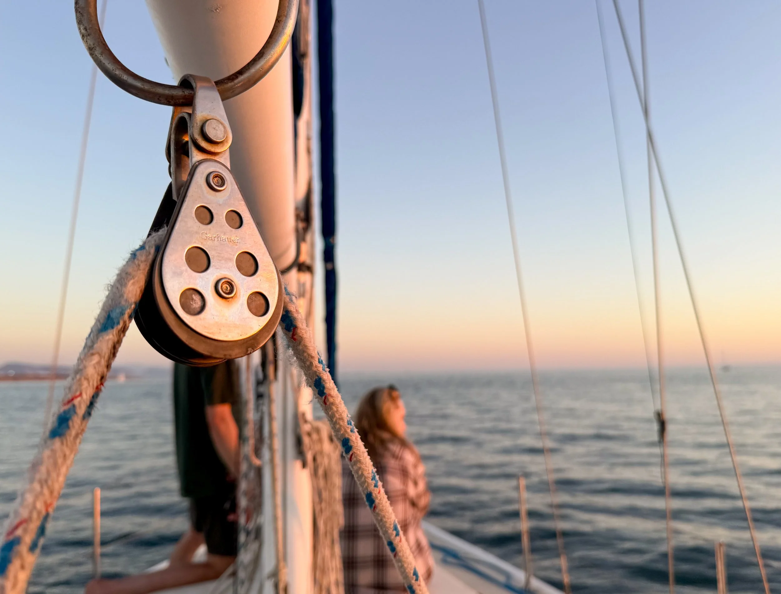 Close-up of a boat's rigging and pulley system on the water during sunset, with a woman with long hair in the background and blurred sailboats on the horizon.