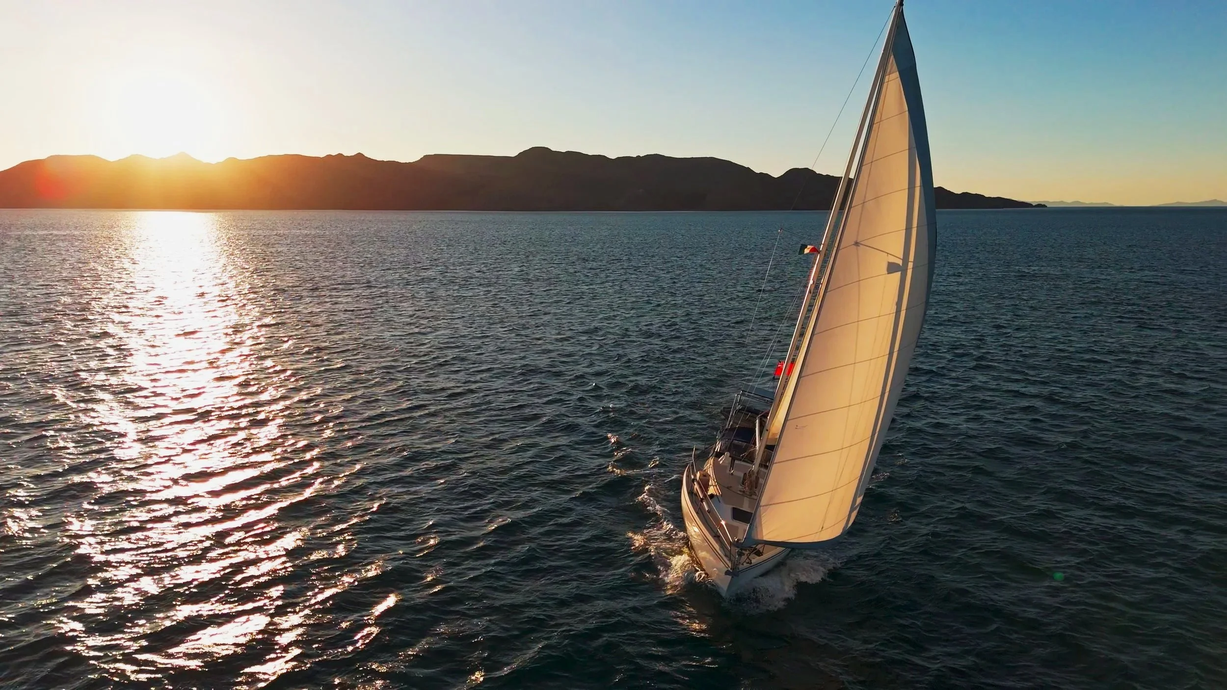 A sailboat with white sails sailing on calm water during sunset with mountains in the background.
