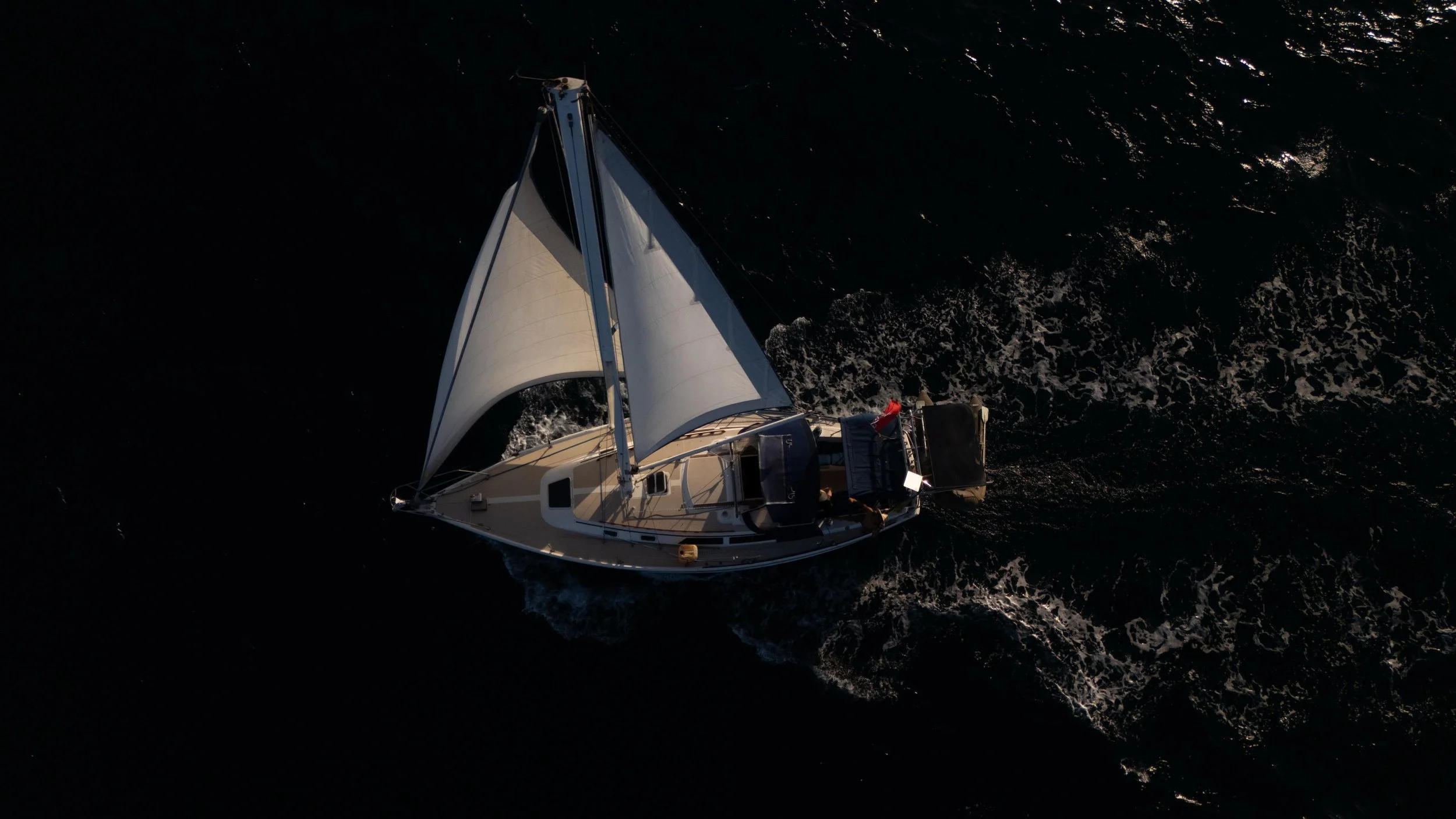 A sailboat with white sails sailing on dark ocean water, viewed from above.