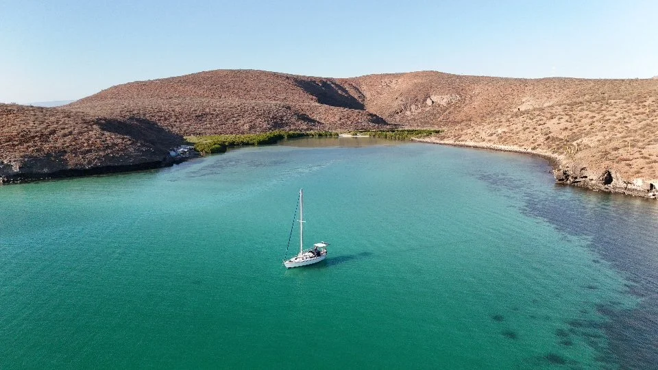A sailboat in turquoise water near brown, arid hills under a clear blue sky.