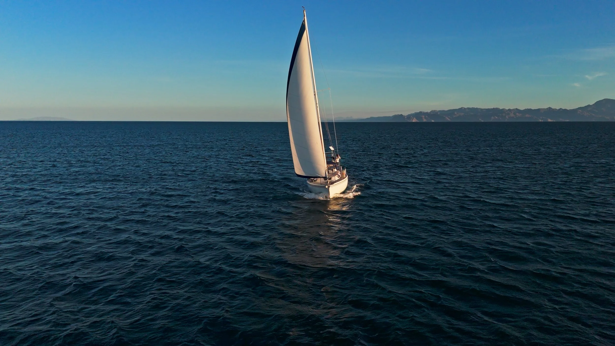 A sailboat with white sails sailing on a calm blue ocean with mountainous land in the background under a clear sky.
