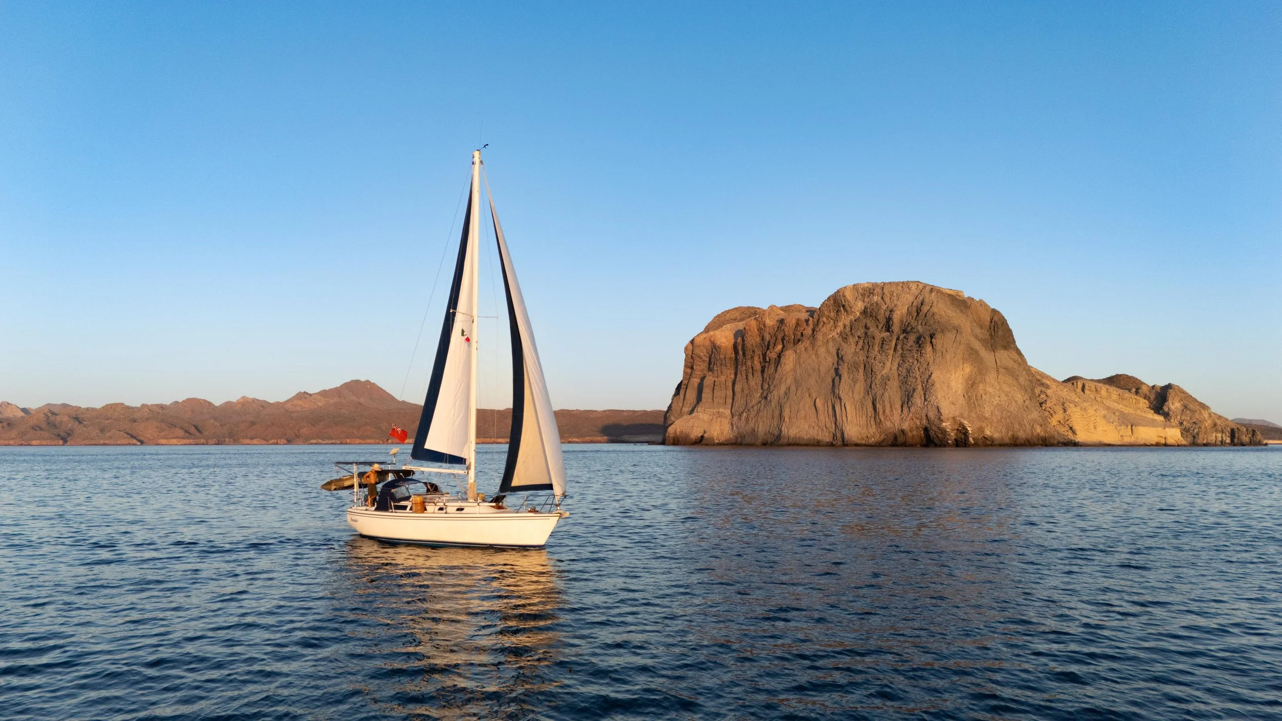 A sailboat with white sails on calm water near a large rocky island during sunset.