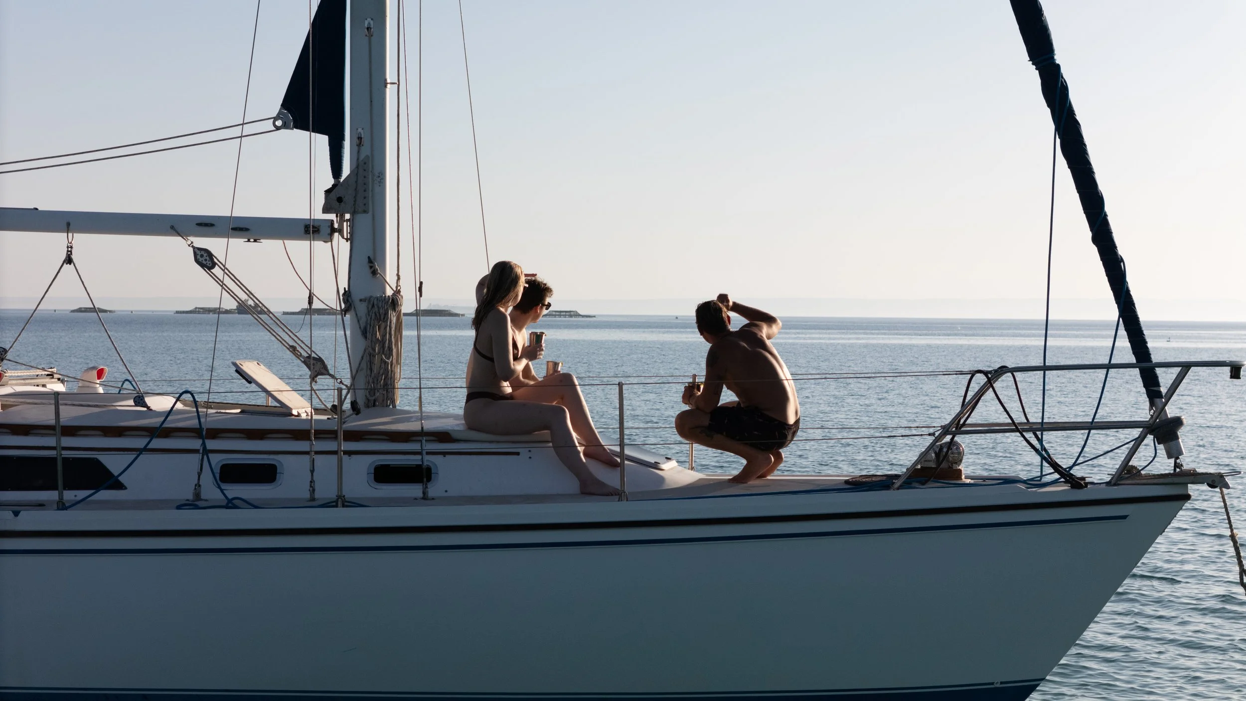 Three people relaxing on a sailboat, with two women sitting and a man squatting, all enjoying drinks while looking at the water and distant islands on a sunny day.