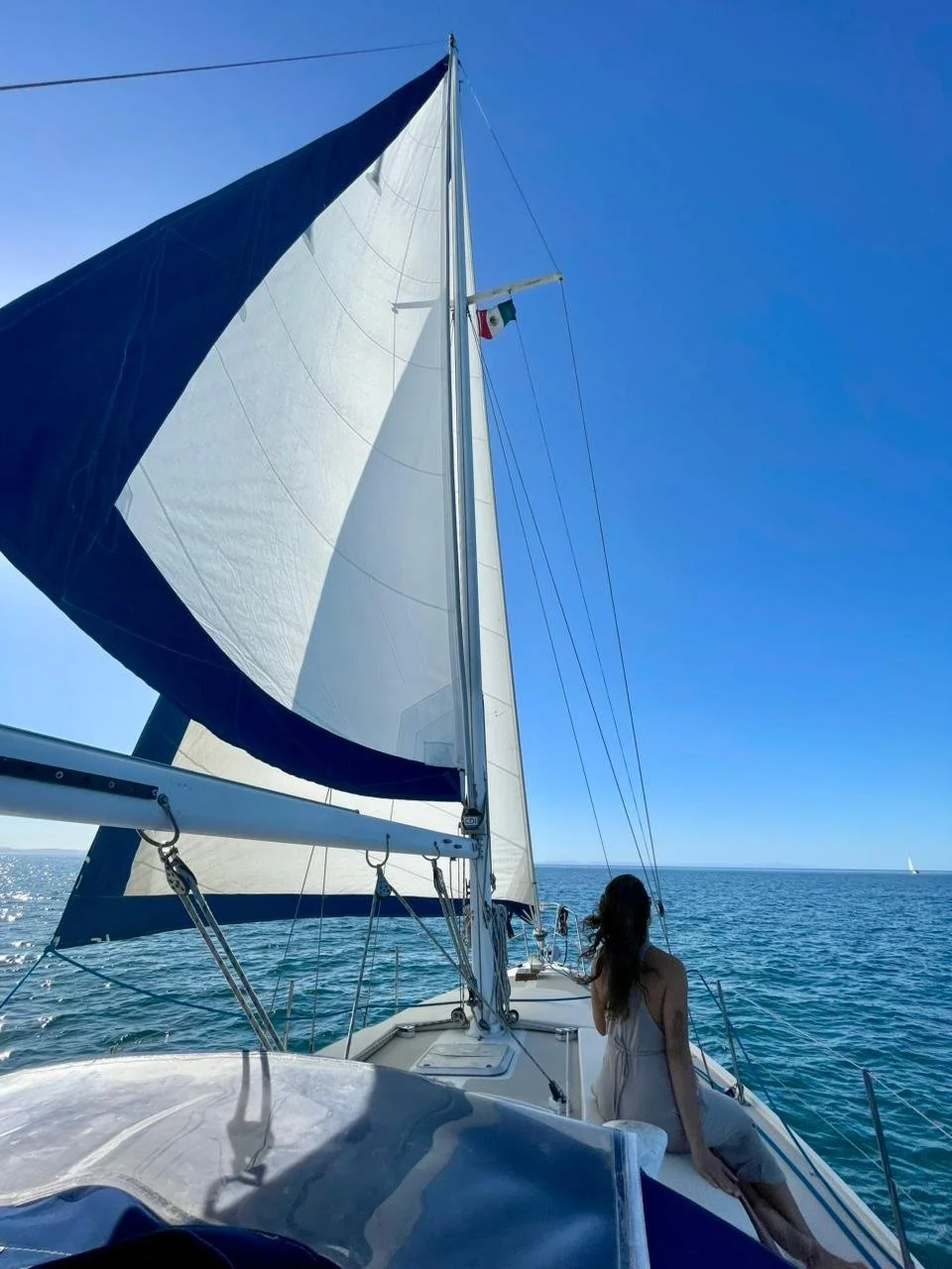 A woman in a white dress sitting on the bow of a sailboat with sails unfurled, on the open water under a clear blue sky, with a small flag near the mast.