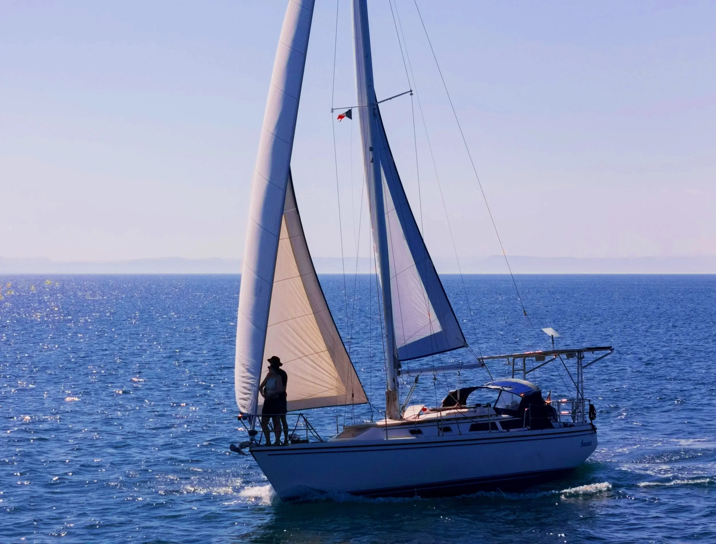A sailboat on the open water with two people on deck, blue sky, and distant land on the horizon.