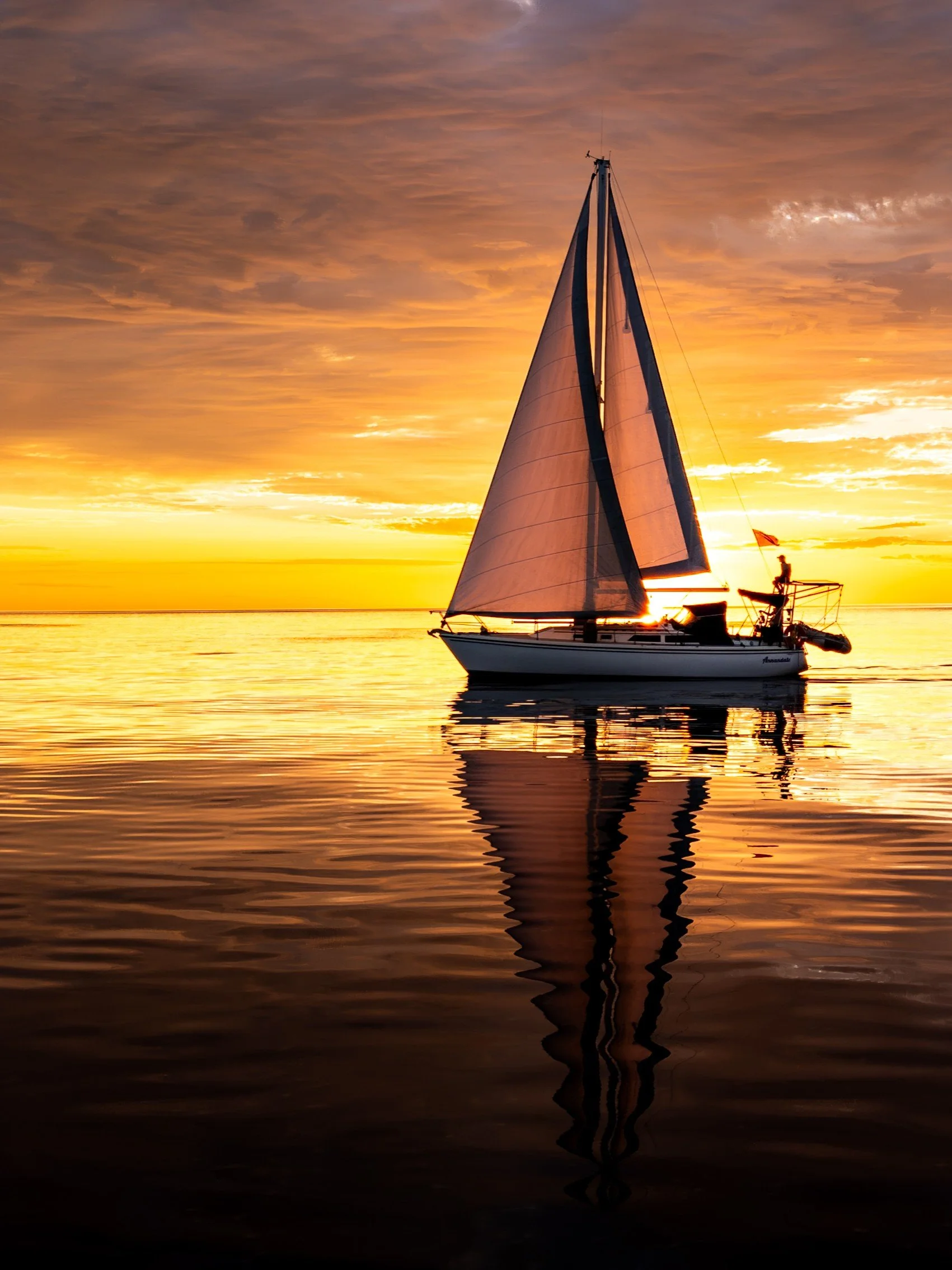 A sailboat on calm water during a vibrant sunset with orange and yellow hues and dramatic clouds in the sky.