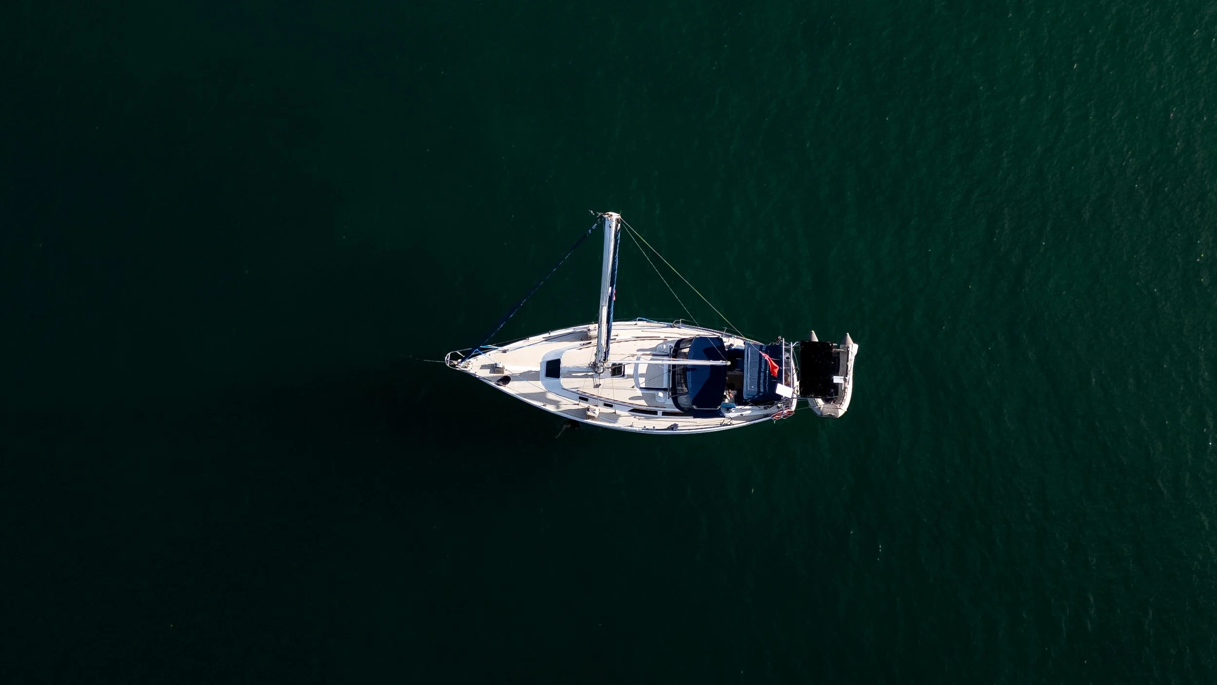 An aerial view of a white sailboat with a mast, floating alone on dark green water.