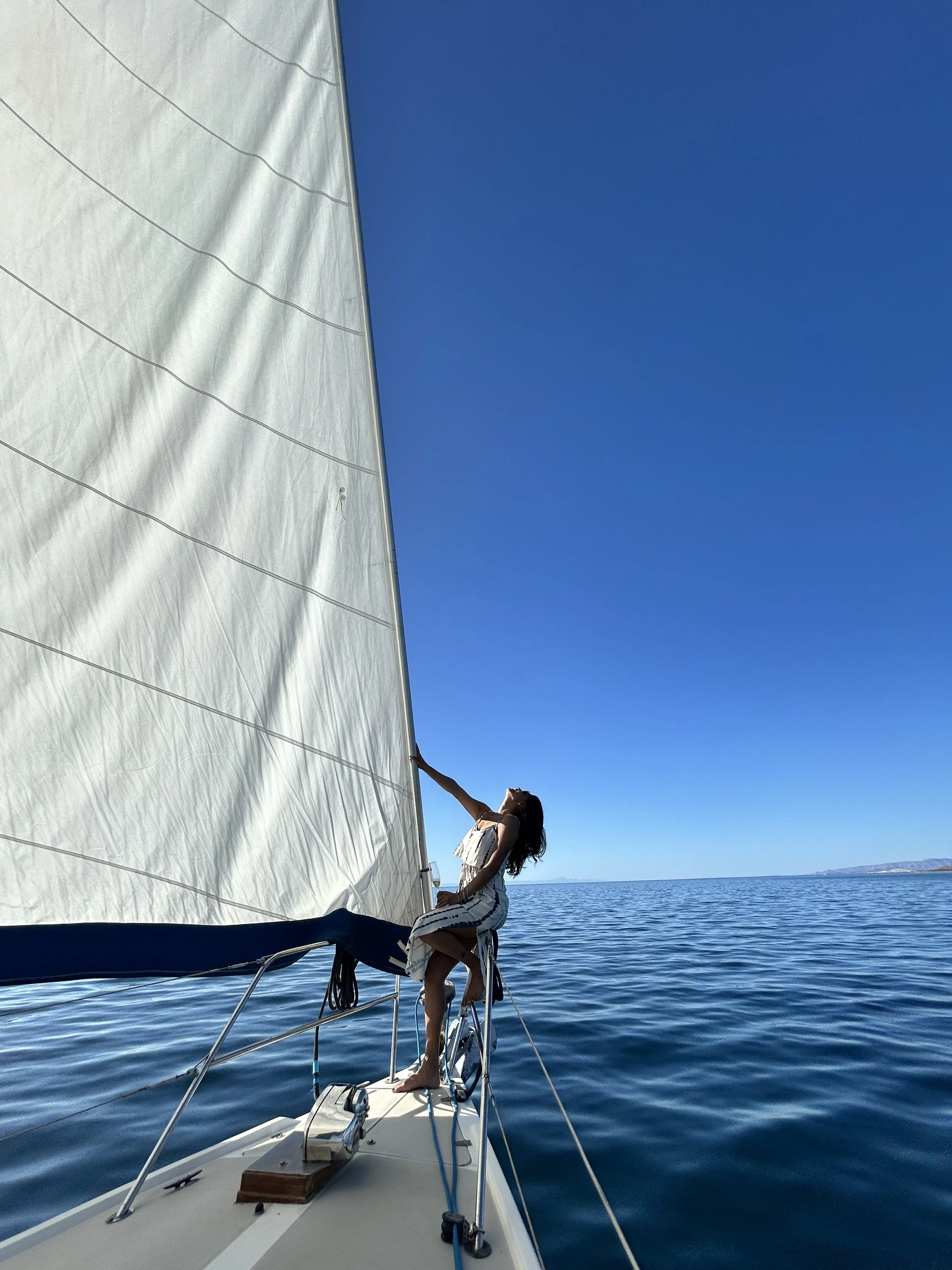 Woman in a white dress sitting on the bow of a sailboat, holding onto the sail, with a clear blue sky and calm sea in the background.