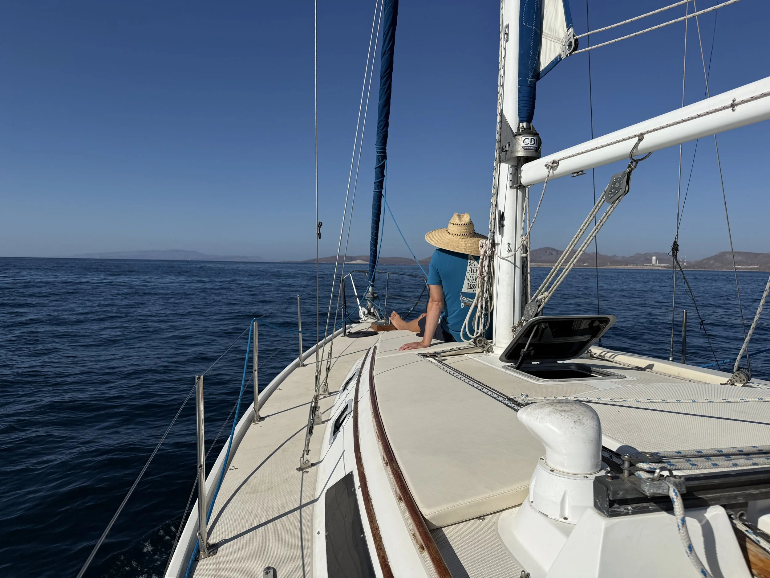 A person sitting on the deck of a sailboat wearing a straw hat, with a view of calm ocean and distant land across the horizon.