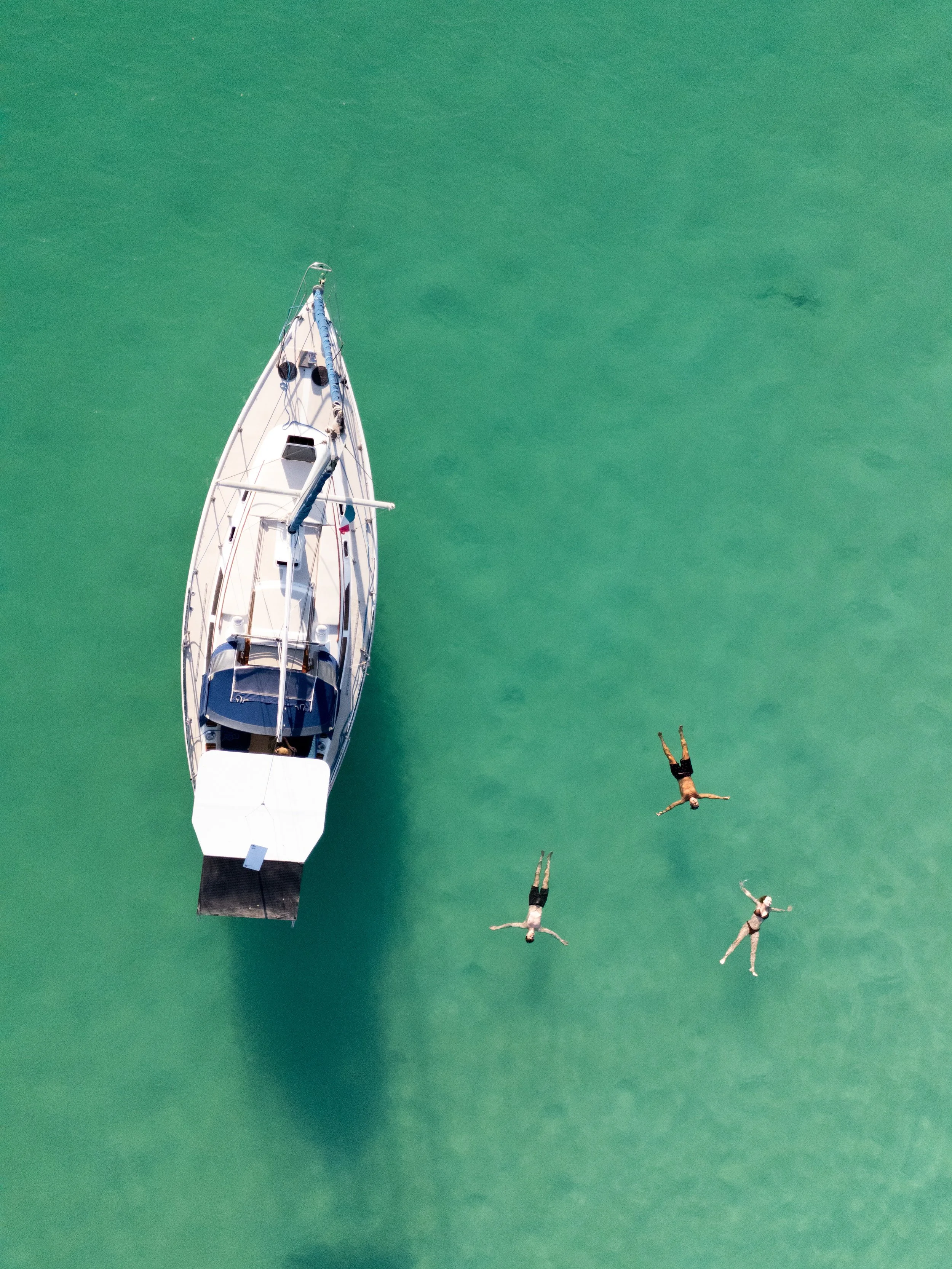 Drone view of a yacht floating on green water with three people lying on their backs nearby, all wearing swimsuits.