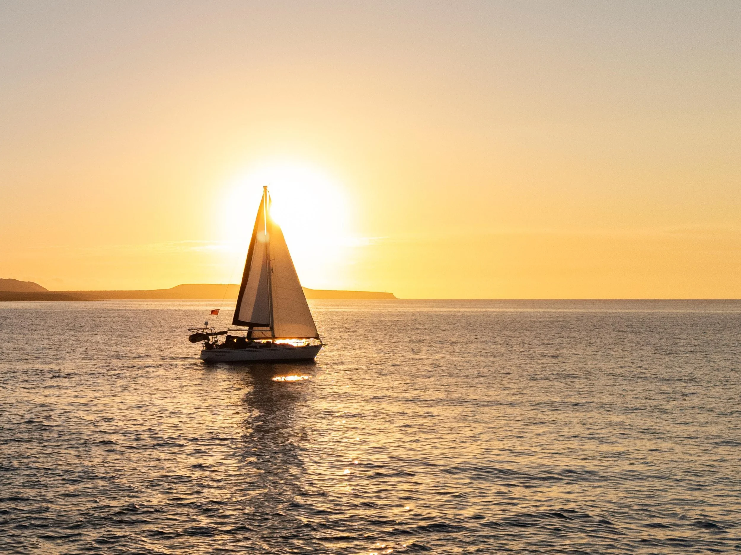 A sailboat on calm water during sunset with the sun low in the sky near the horizon.